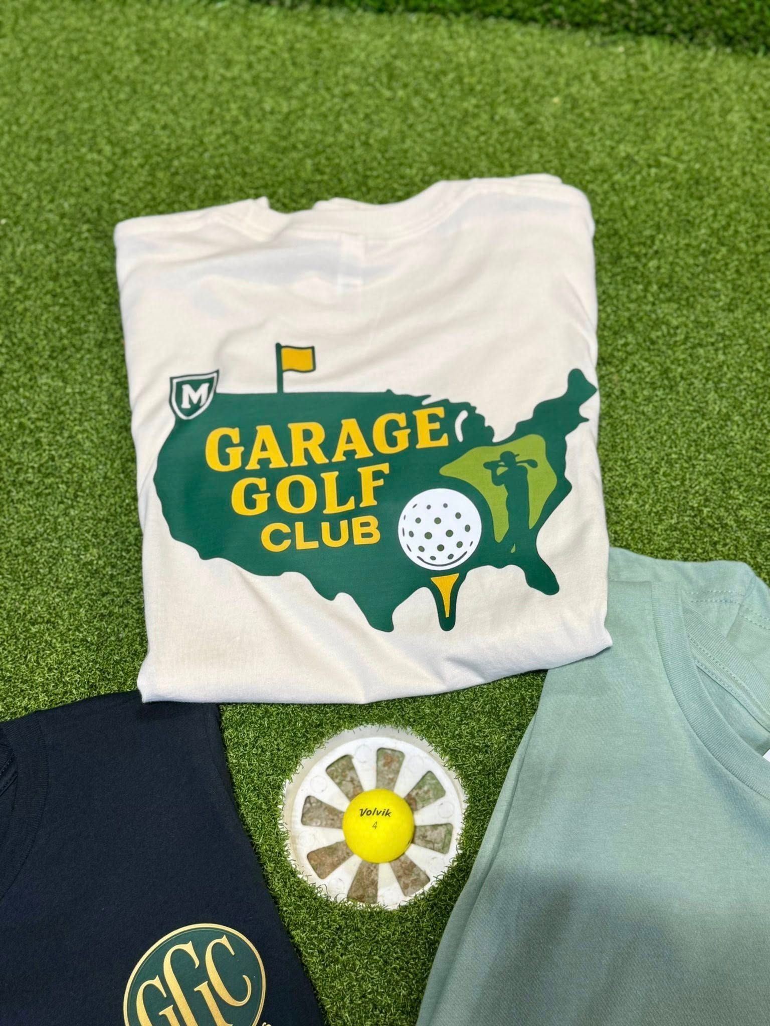 Cream Garage Golf Club t-shirt with logo, golf ball marker, and two other shirts on green turf.