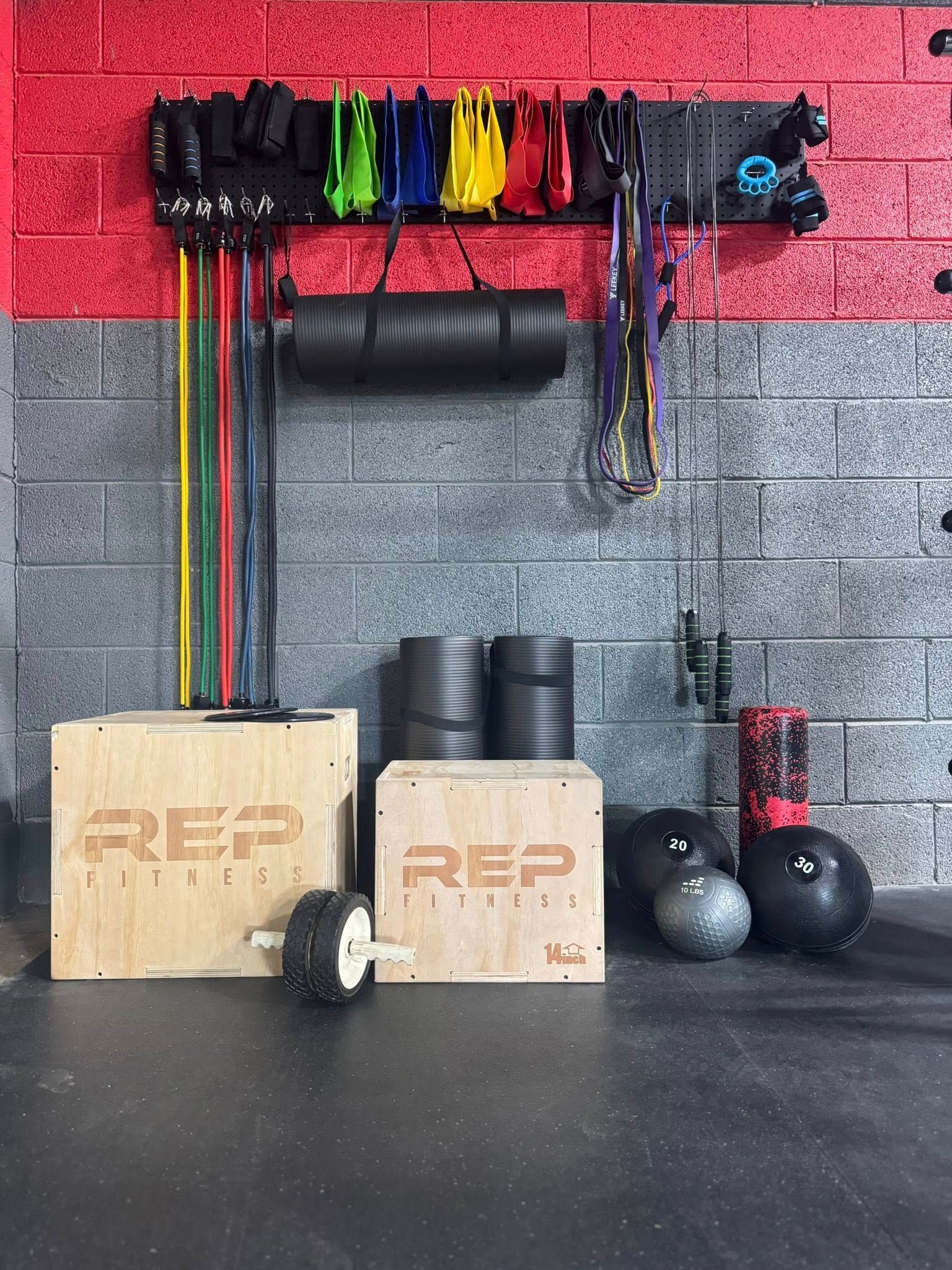 Gym equipment: resistance bands, boxes, medicine balls, and foam rollers on a black floor against gray and red walls.