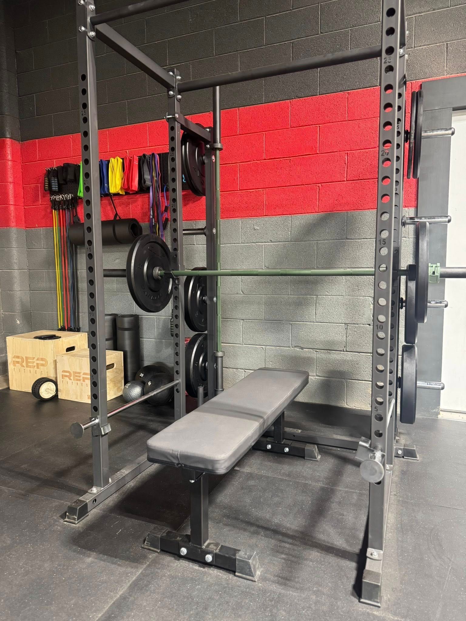 Weightlifting rack with bench, barbell loaded with weights, plates stored on the rack. Red and gray gym interior.