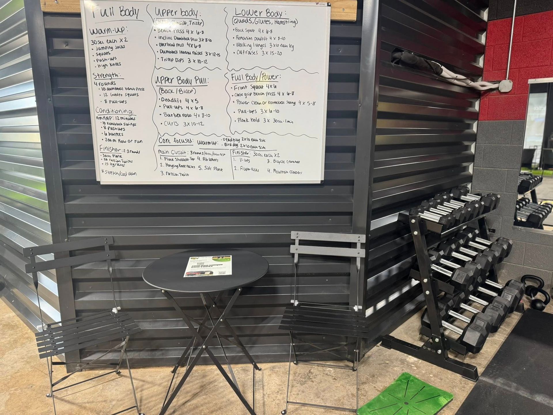 Black outdoor fitness area with a whiteboard, weights, table, and chairs.