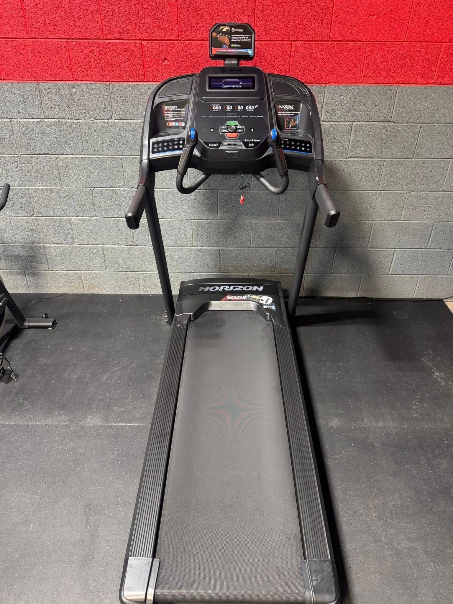 Treadmill with a black running surface, digital display, and dark frame, positioned in front of a red and grey wall.