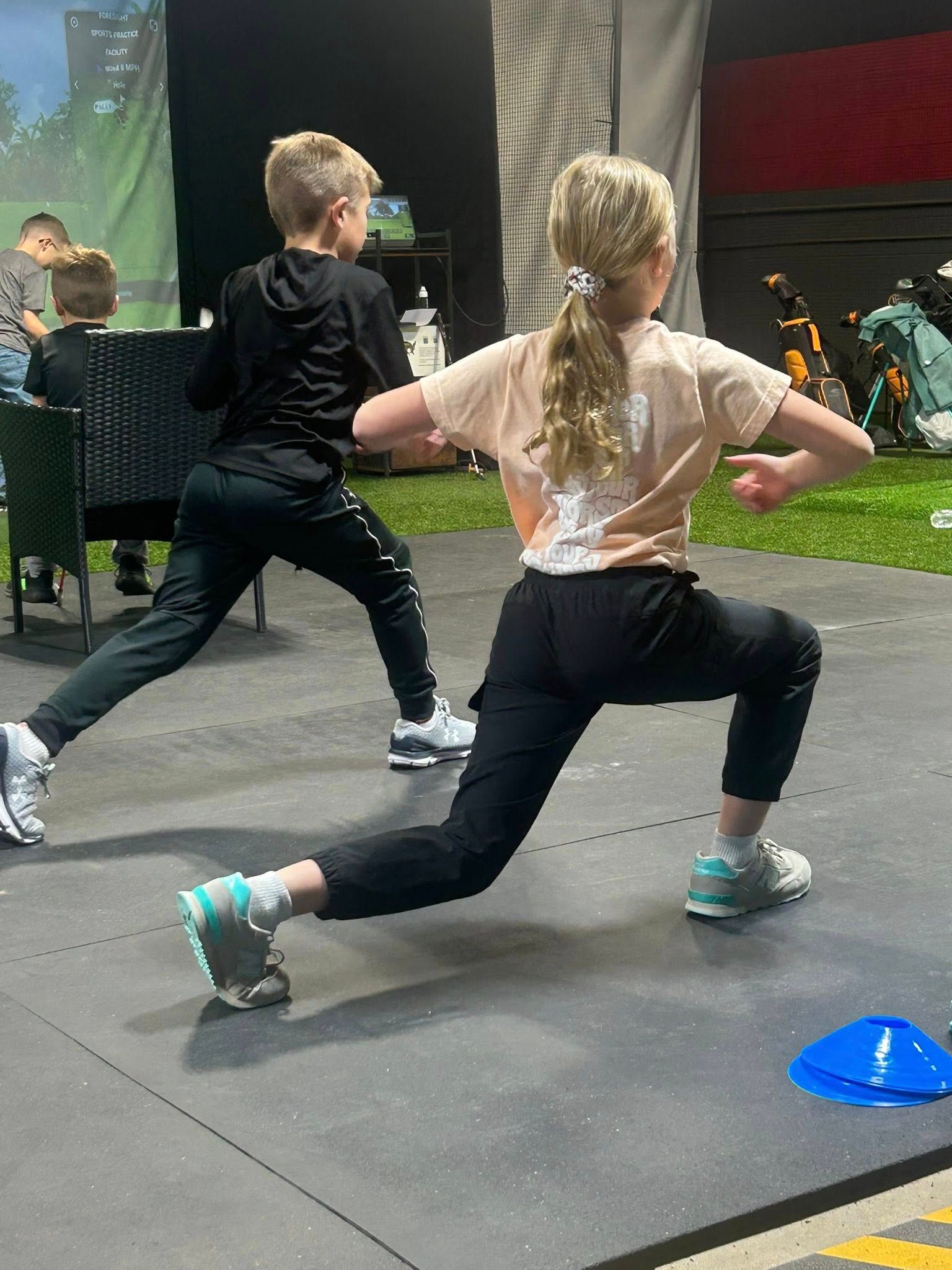 Two children doing lunges on a gray mat. One child has blond hair in a ponytail, arms out. The other child is wearing a black hoodie.