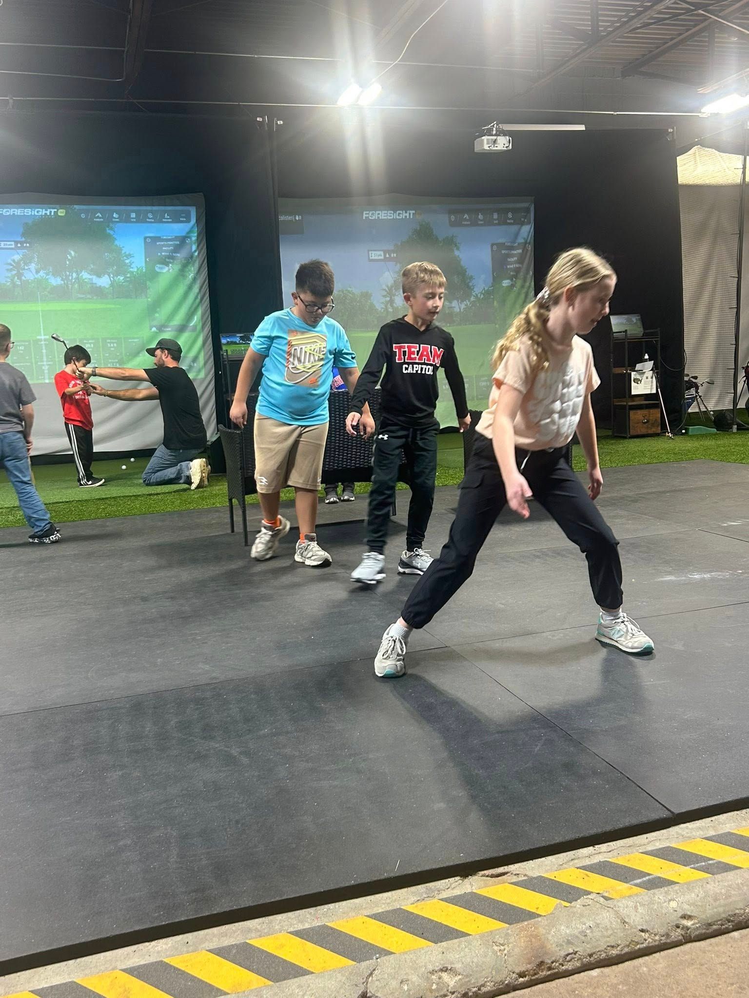 Children practicing golf in an indoor facility. Two golf simulators are visible in the background.
