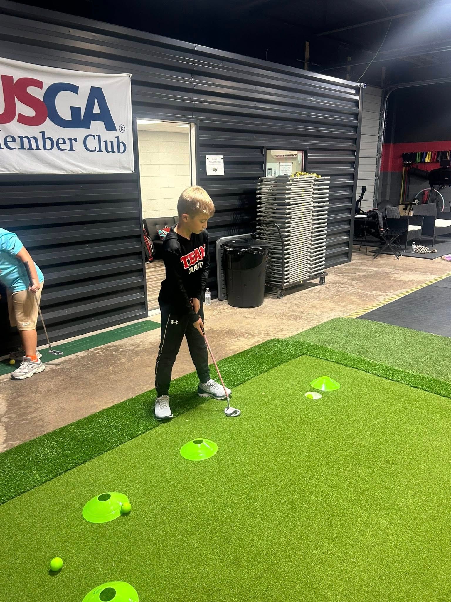 A young person playing golf indoors on a green putting surface, aiming for a hole.