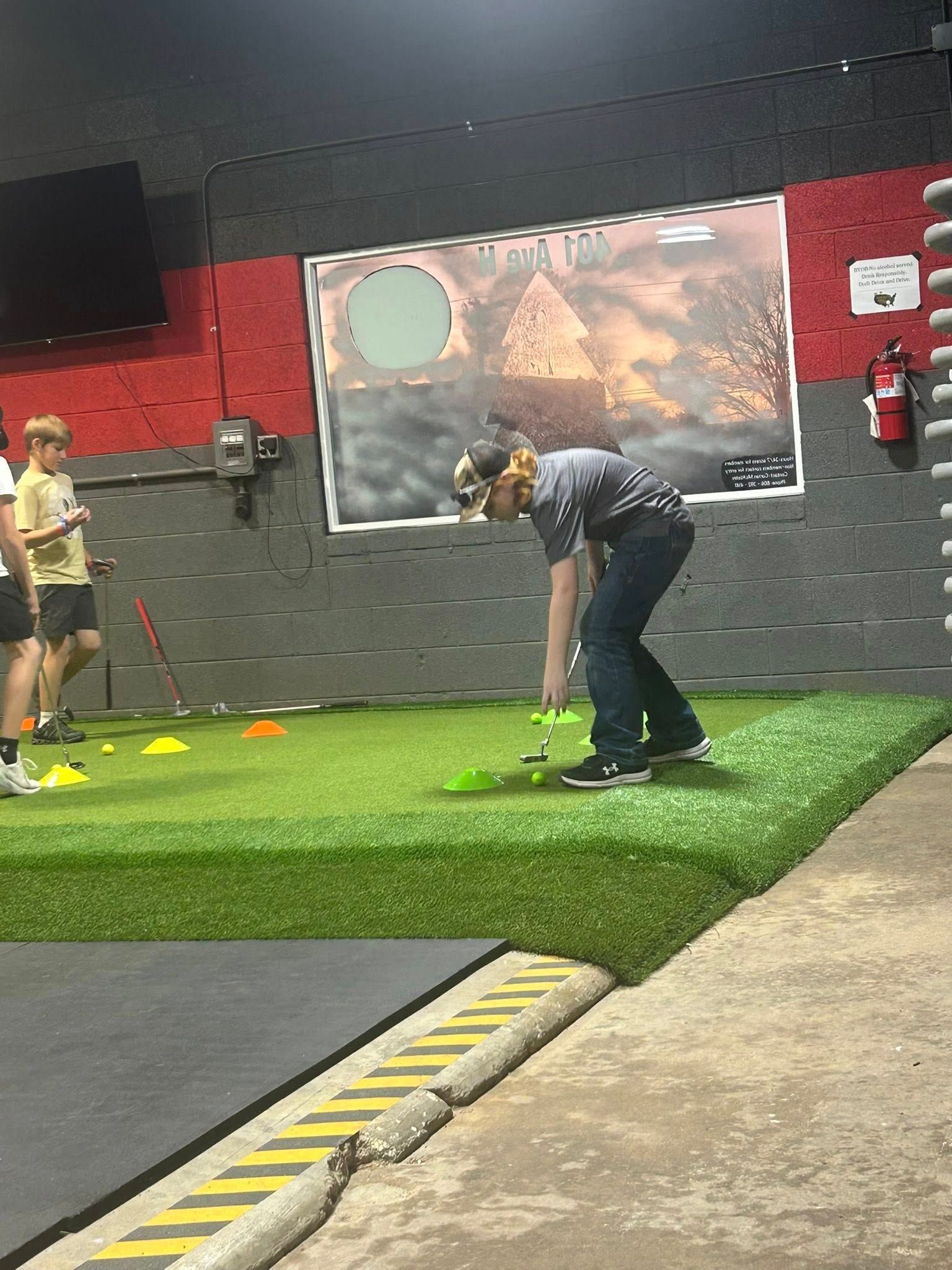 Person playing mini golf indoors, on a turf-covered platform. Other people watch.