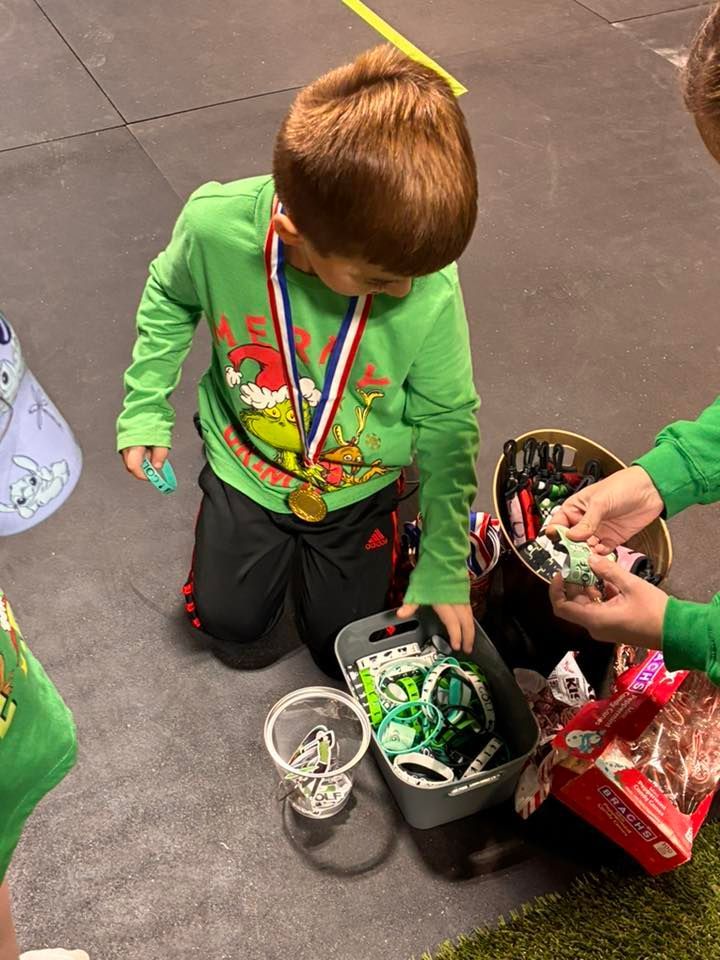 Boy kneeling, selecting items from a bin. He wears a medal, Grinch shirt. Other children nearby, gifts, and a green mat.
