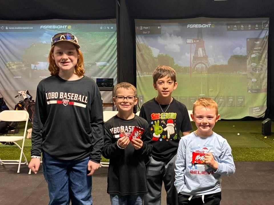 Four young boys smiling in a golf simulator room, two holding prizes, screens in background.
