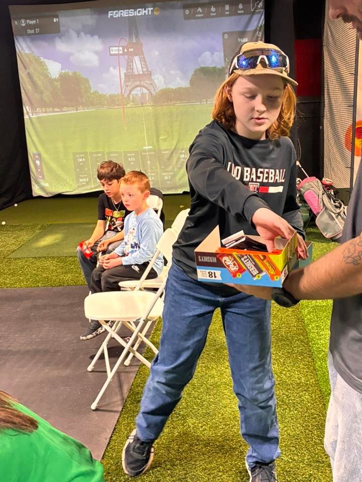 A person handing snacks to another person in a golf simulator, two children watch in the background.