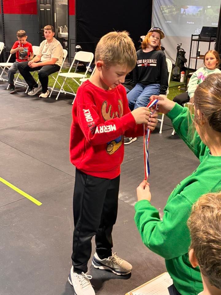 Boy in red sweater receiving a medal. Others watch. Indoor setting.