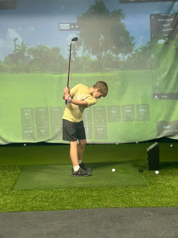 Young person swings a golf club indoors toward a screen, on a turf mat.