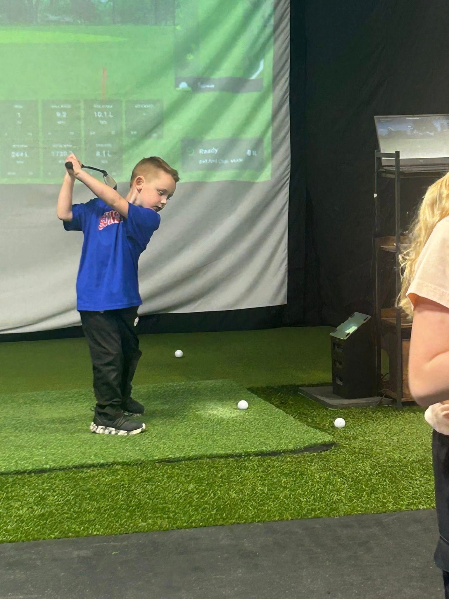 Boy swinging a golf club indoors at a simulator, green turf, wearing a blue shirt.