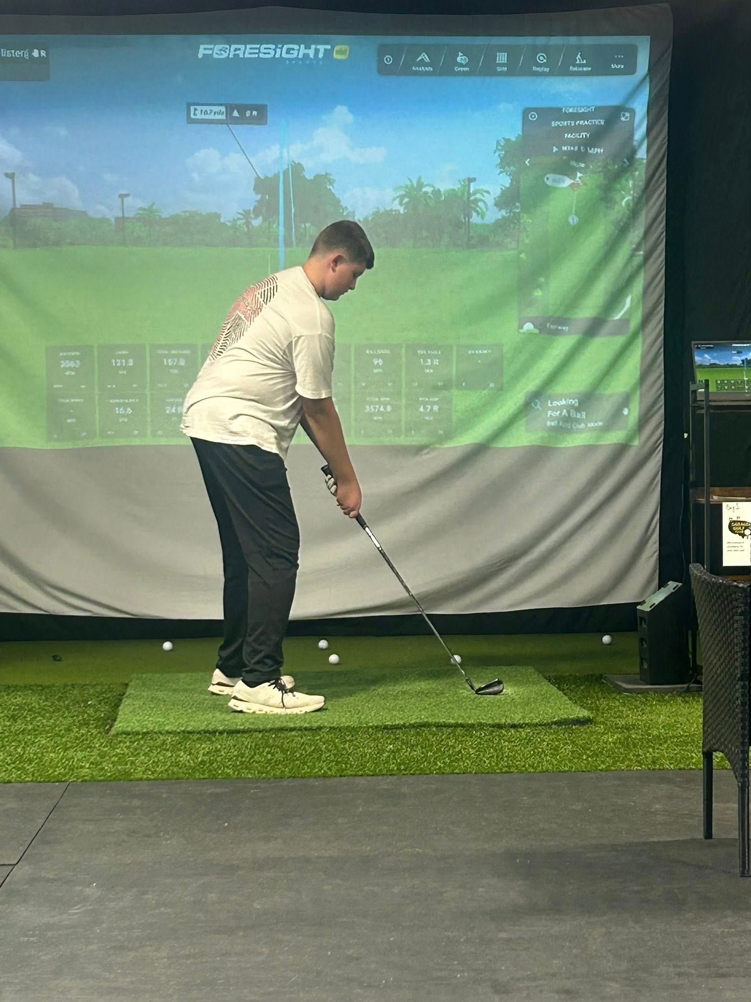 Person in a golf simulator, preparing to swing. He's holding a club, with screen displaying a course.