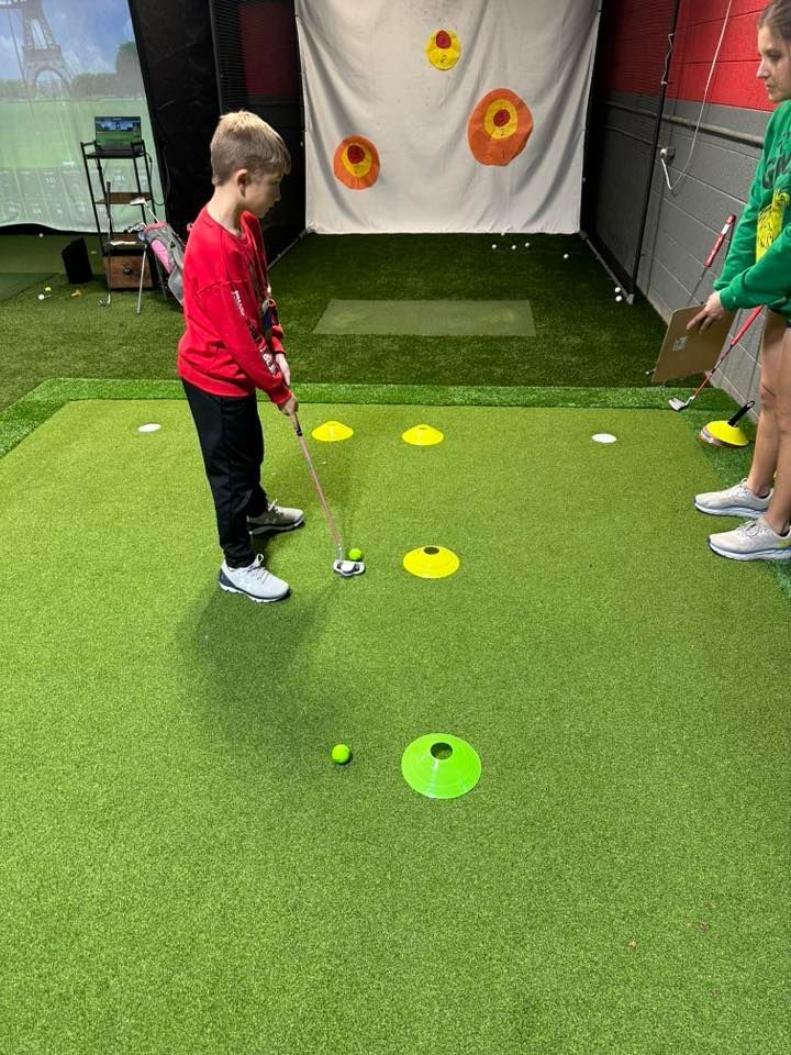 Boy in red shirt putting golf ball on a green, with cones. Another person holds a clipboard.