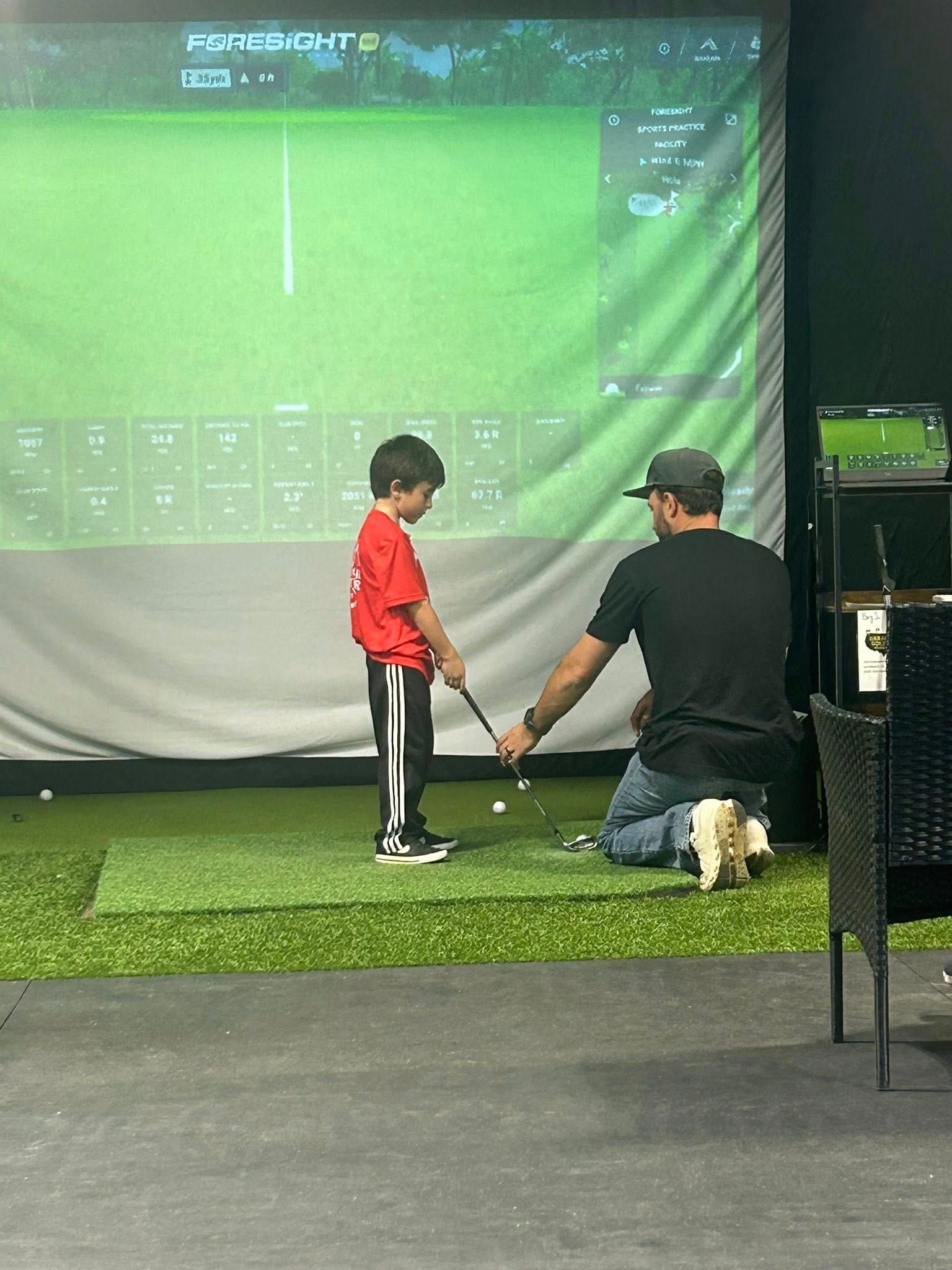 A child in red shirt practices golf swing indoors with an instructor, using a golf simulator.