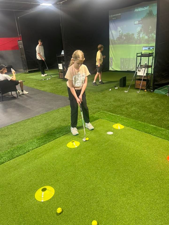 A girl putts a golf ball on an indoor green, other people play golf in the background.