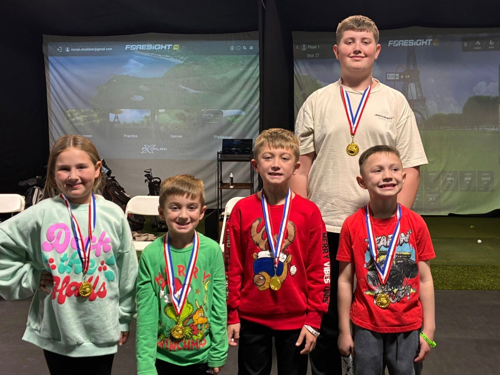 Children wearing medals stand in front of golf simulator screens.