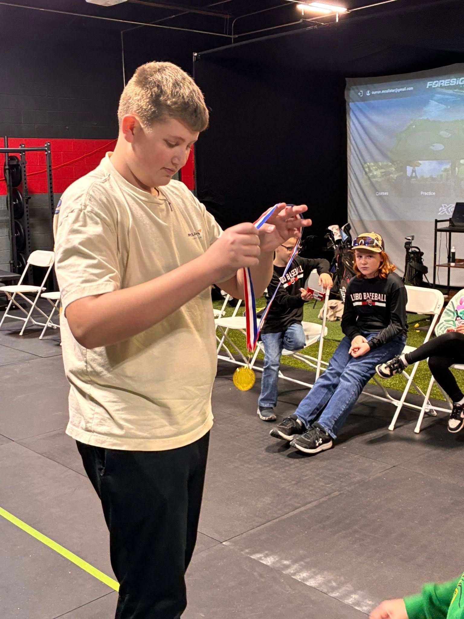 Boy holding a medal, looking at it. Others sit on chairs in a golf simulator setting.
