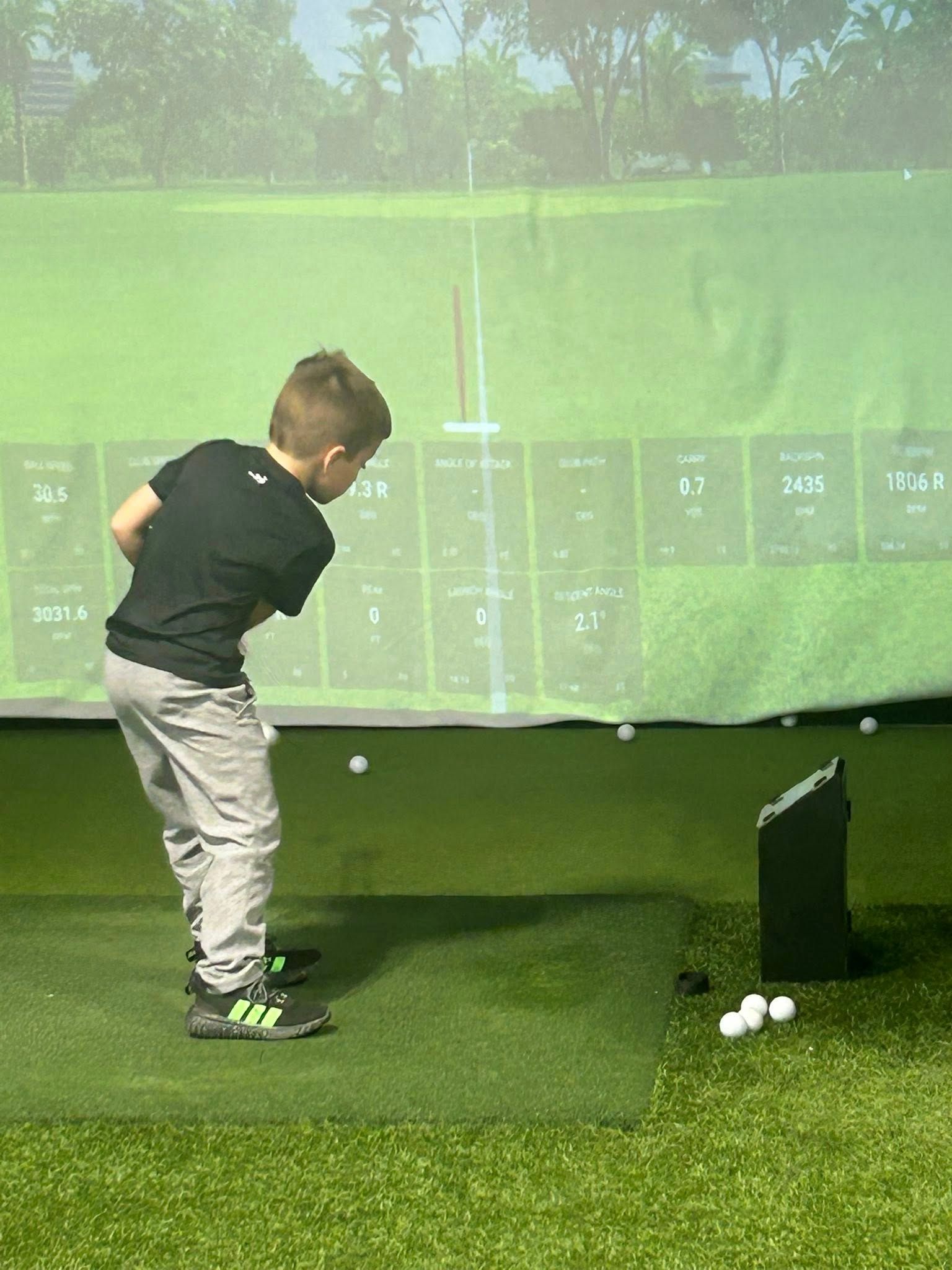 Child in gray pants and black shirt swinging a golf club in a simulator, green turf and screen background.