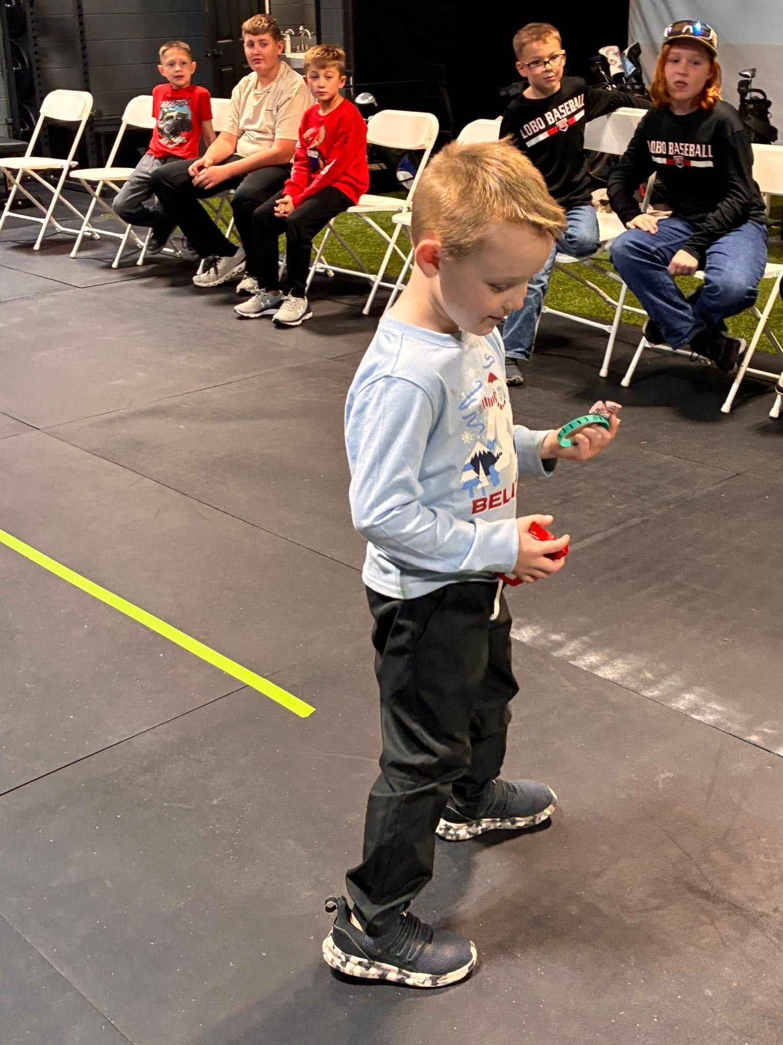 Boy holds a toy, standing on a dark floor with a yellow line. Other children sit on chairs in the background.