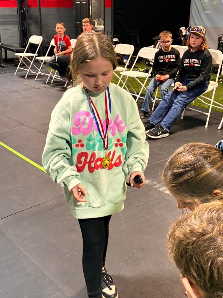 Girl in green sweatshirt with medal stands indoors, looking down. Other kids sit behind her.