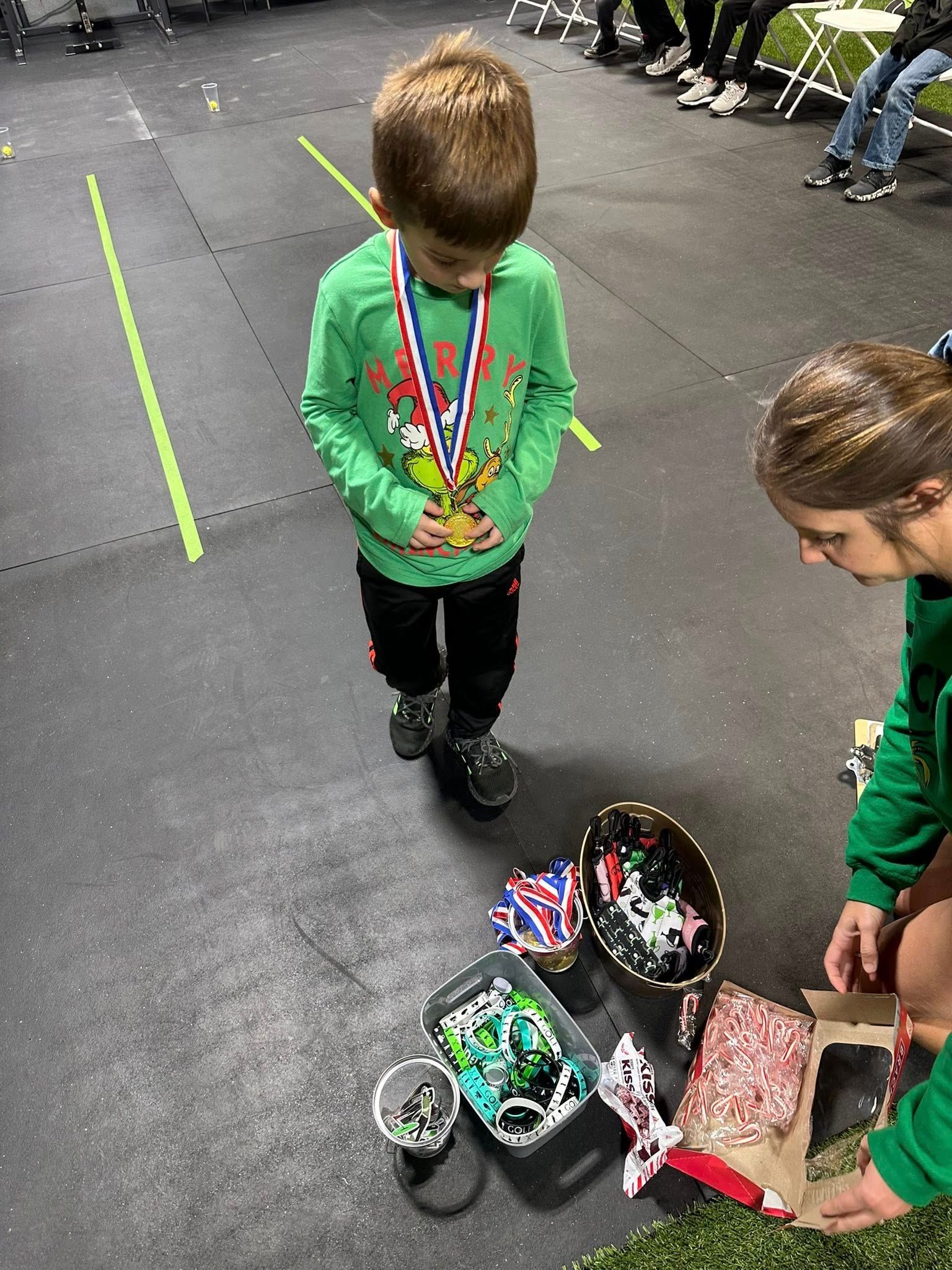 A young boy with a medal stands next to a woman holding treats after an event on a gym floor.