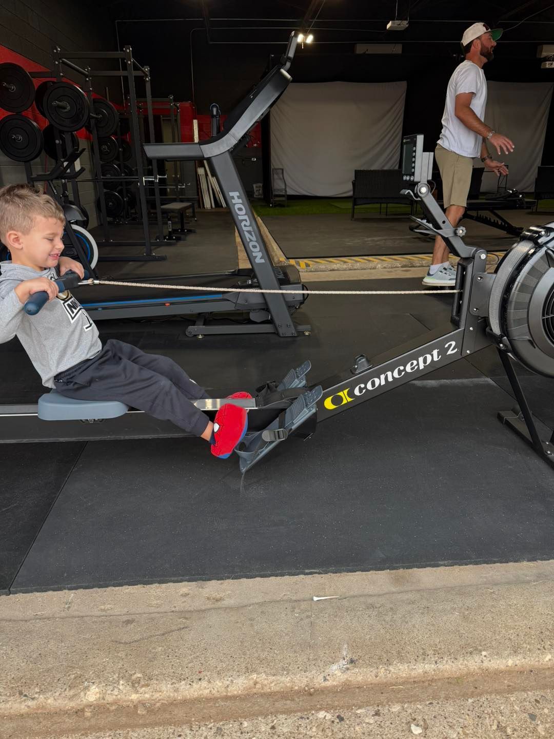 Young child using a Concept 2 rowing machine, smiling. Adult in background. Gym setting.