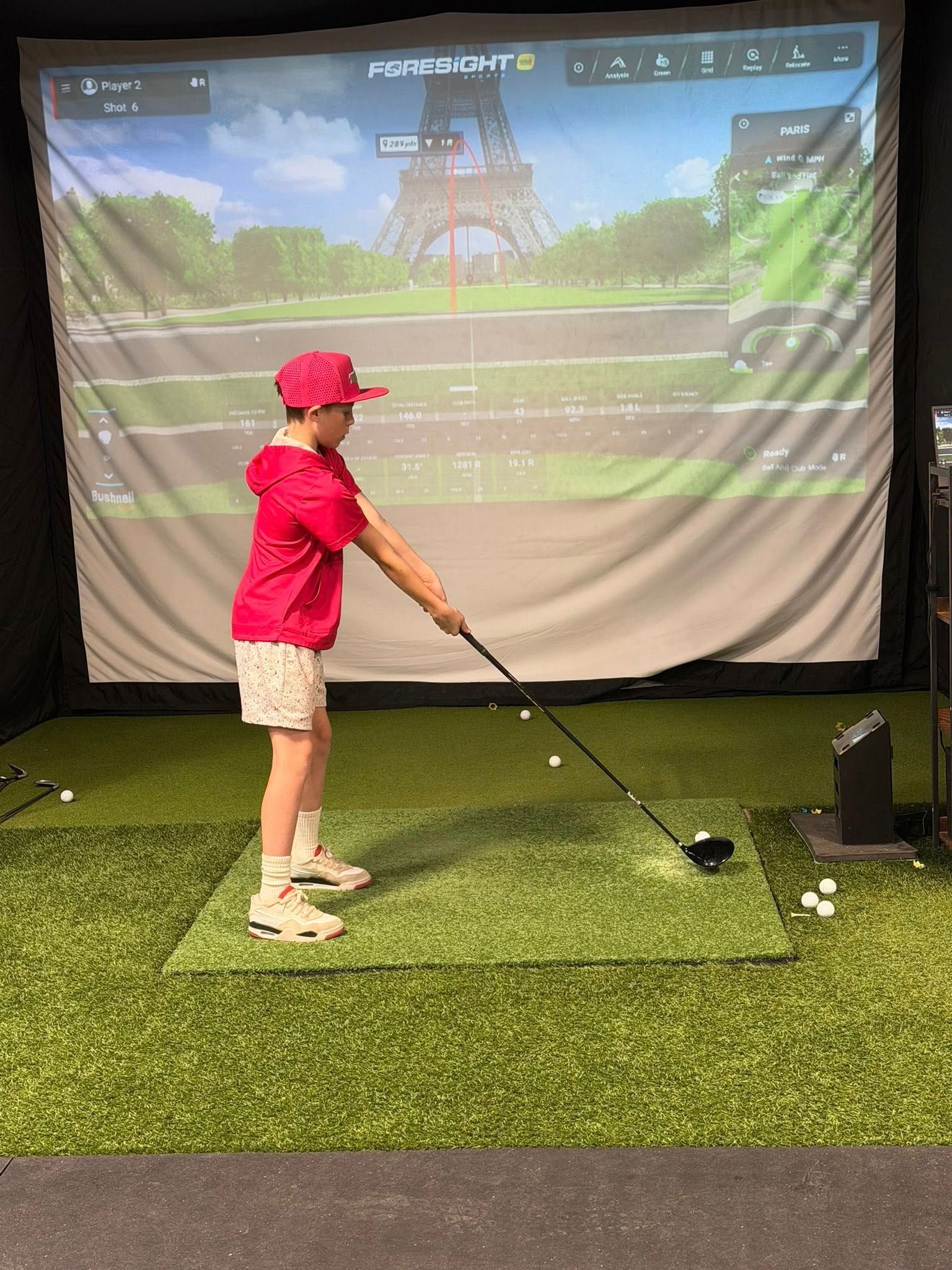 Young person in pink hitting a golf ball in a simulator; Eiffel Tower background.