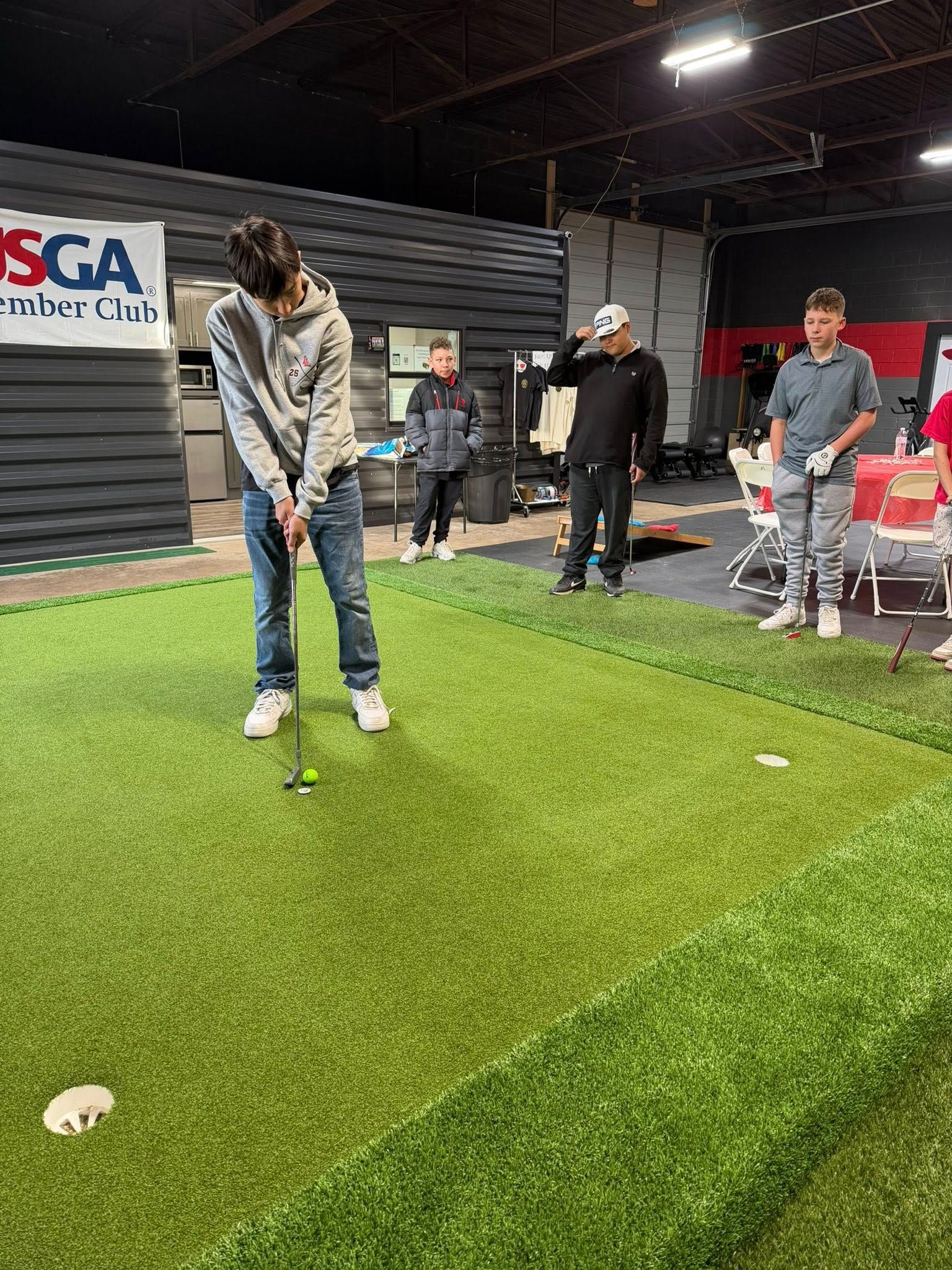 Person putting golf ball on a green with others watching indoors. Green turf, neutral background.