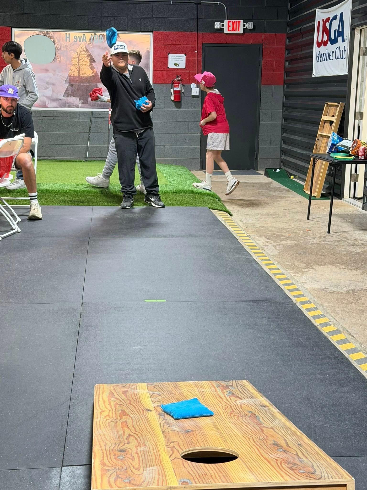 Person tossing a blue beanbag toward a cornhole board in an indoor setting.