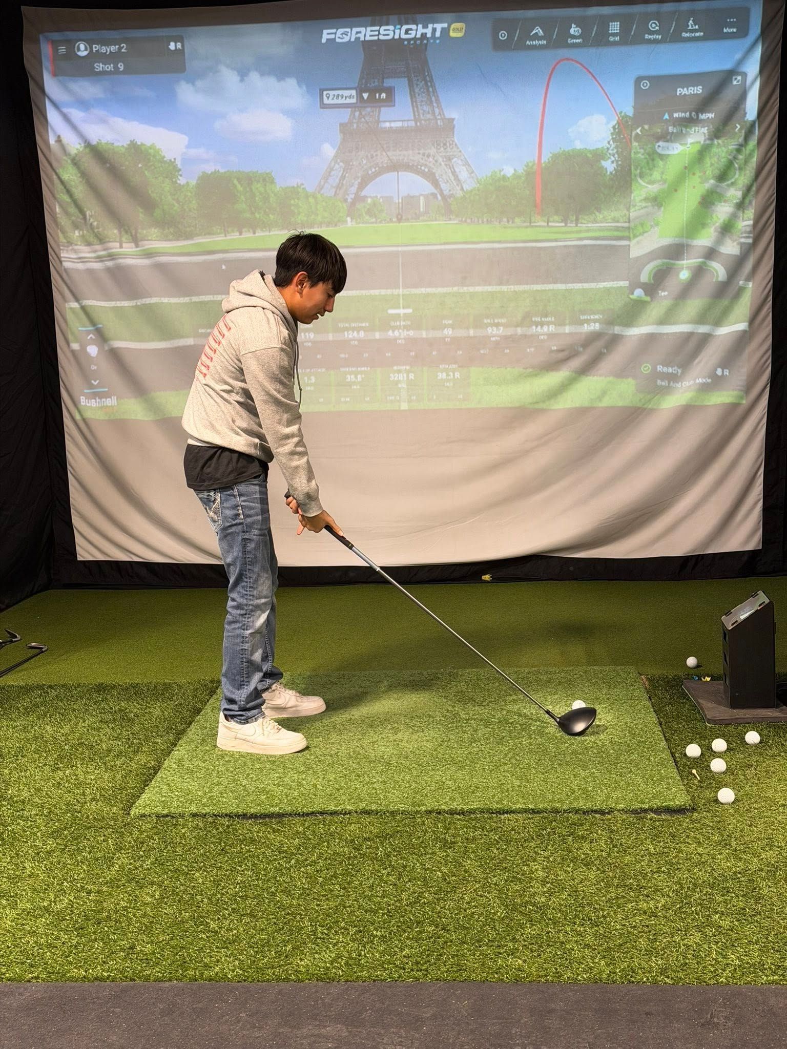Person swinging a golf club in a simulator, aiming towards a screen displaying a golf course with the Eiffel Tower.