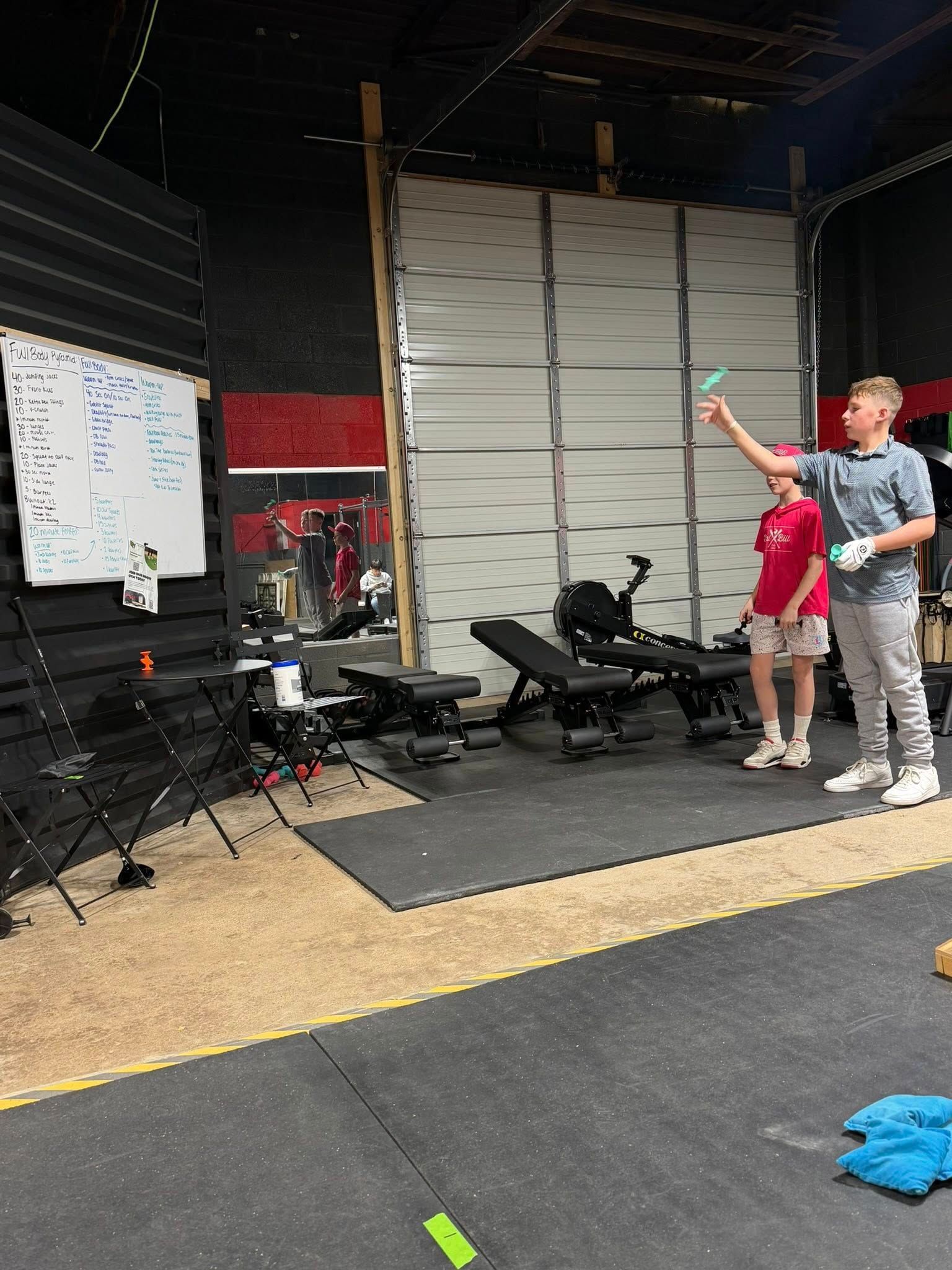 Two boys in a gym throwing foam darts. One aims, the other watches. Whiteboard, weights, and garage door in background.