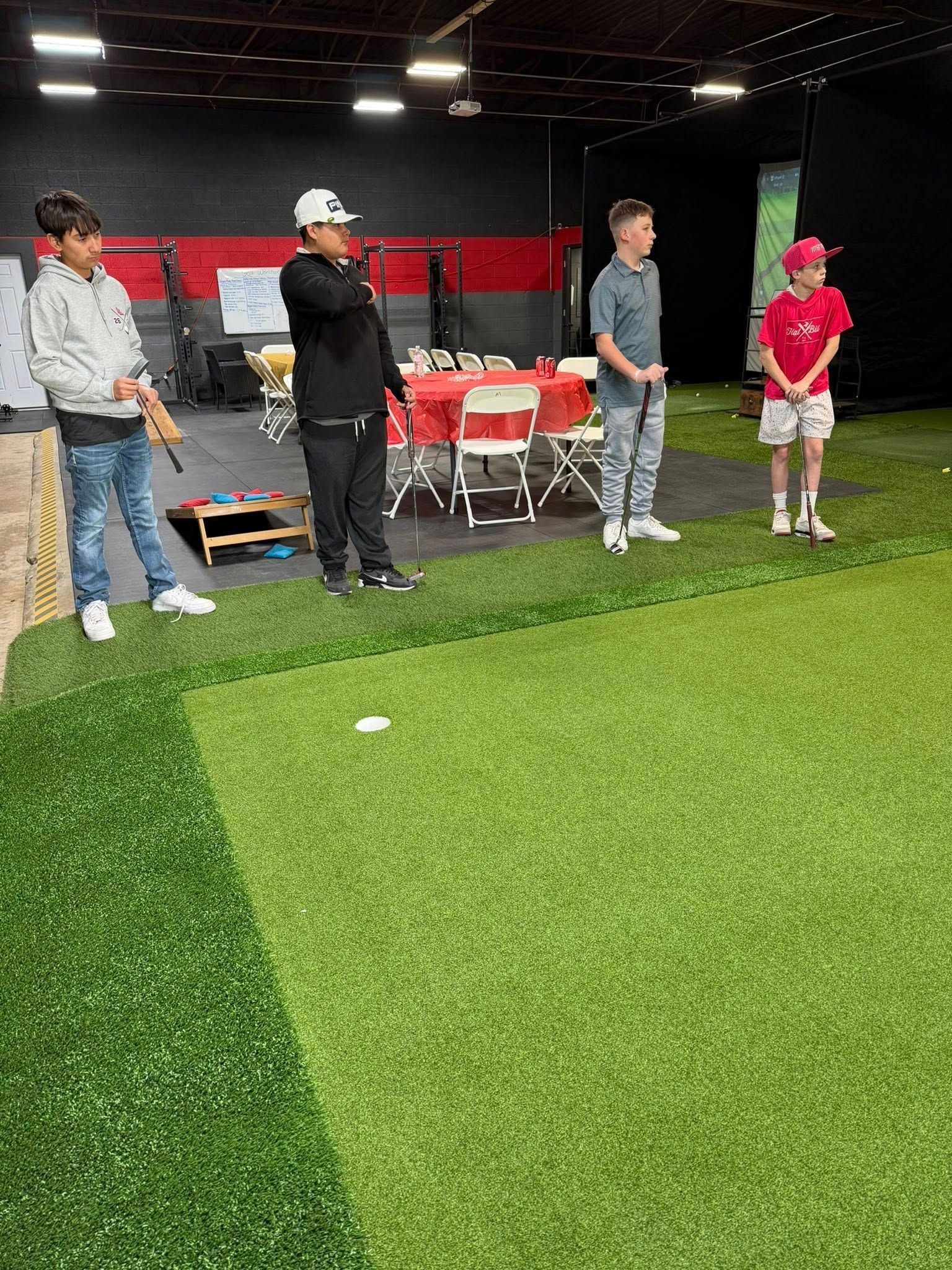 Four people standing on green turf, one with a golf club. Red and white table setup visible in the background.