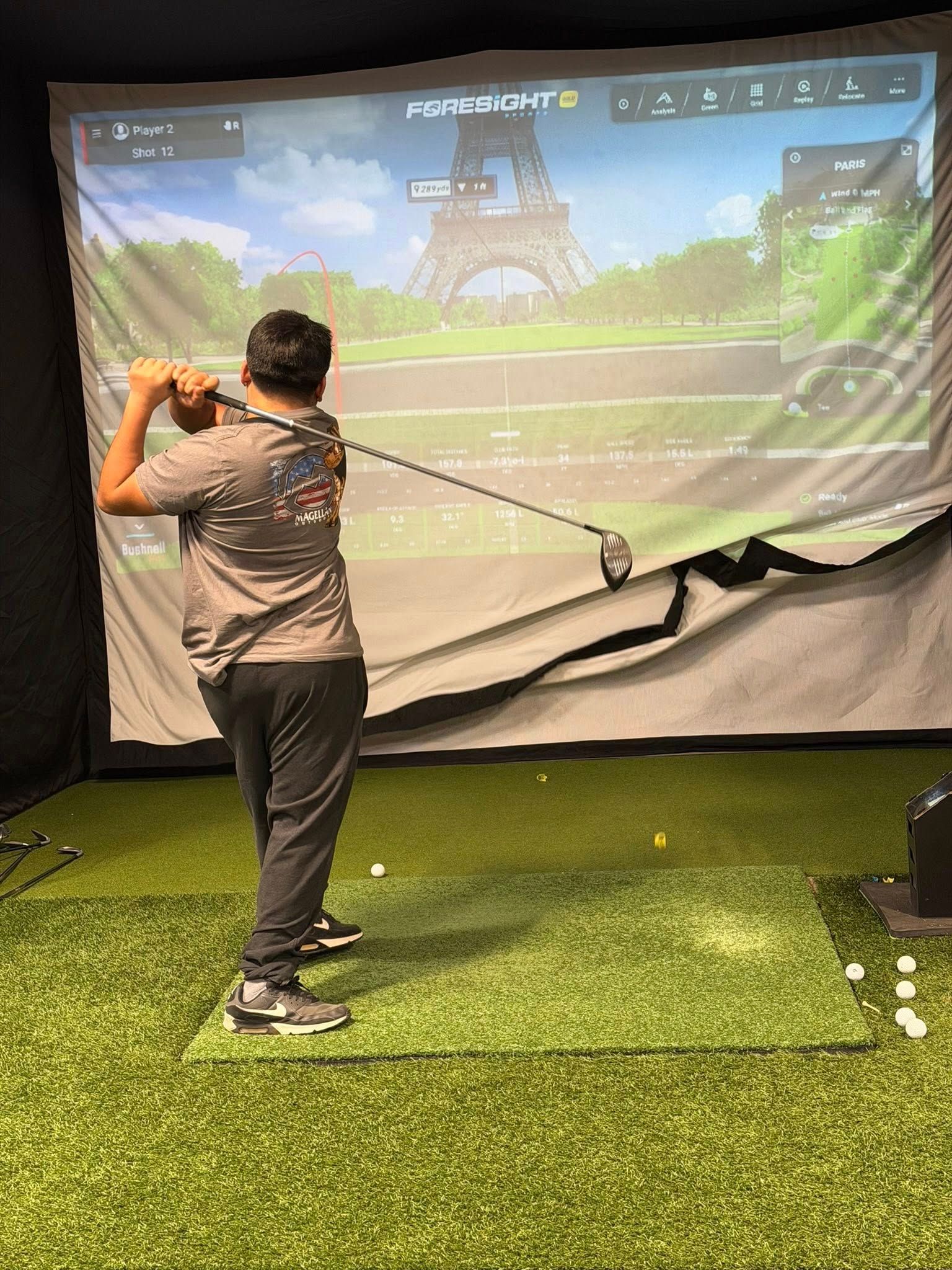 Person swinging a golf club in a simulator with the Eiffel Tower on the screen.
