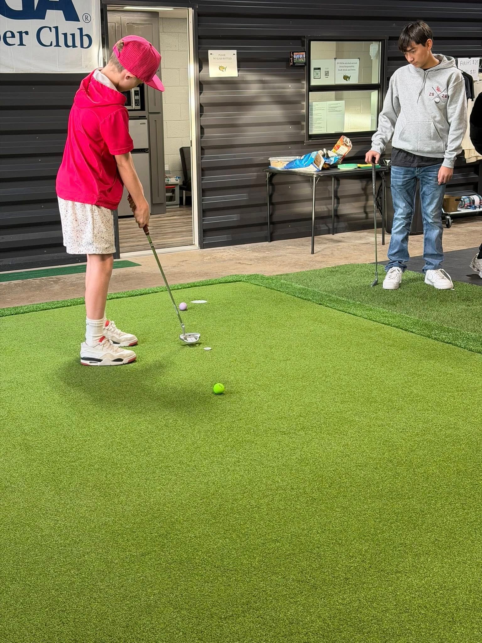Person putting a golf ball on artificial turf, another person watches. Building in background.