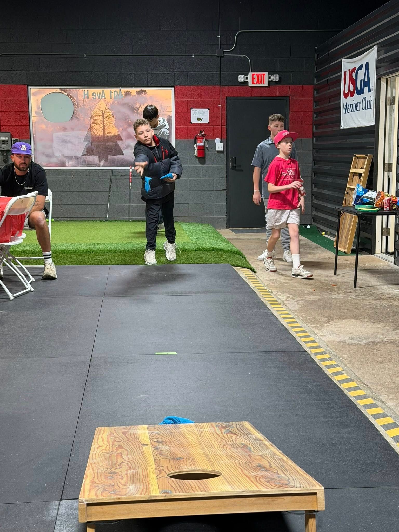 A child throws a beanbag towards a cornhole board indoors. Other people watch.