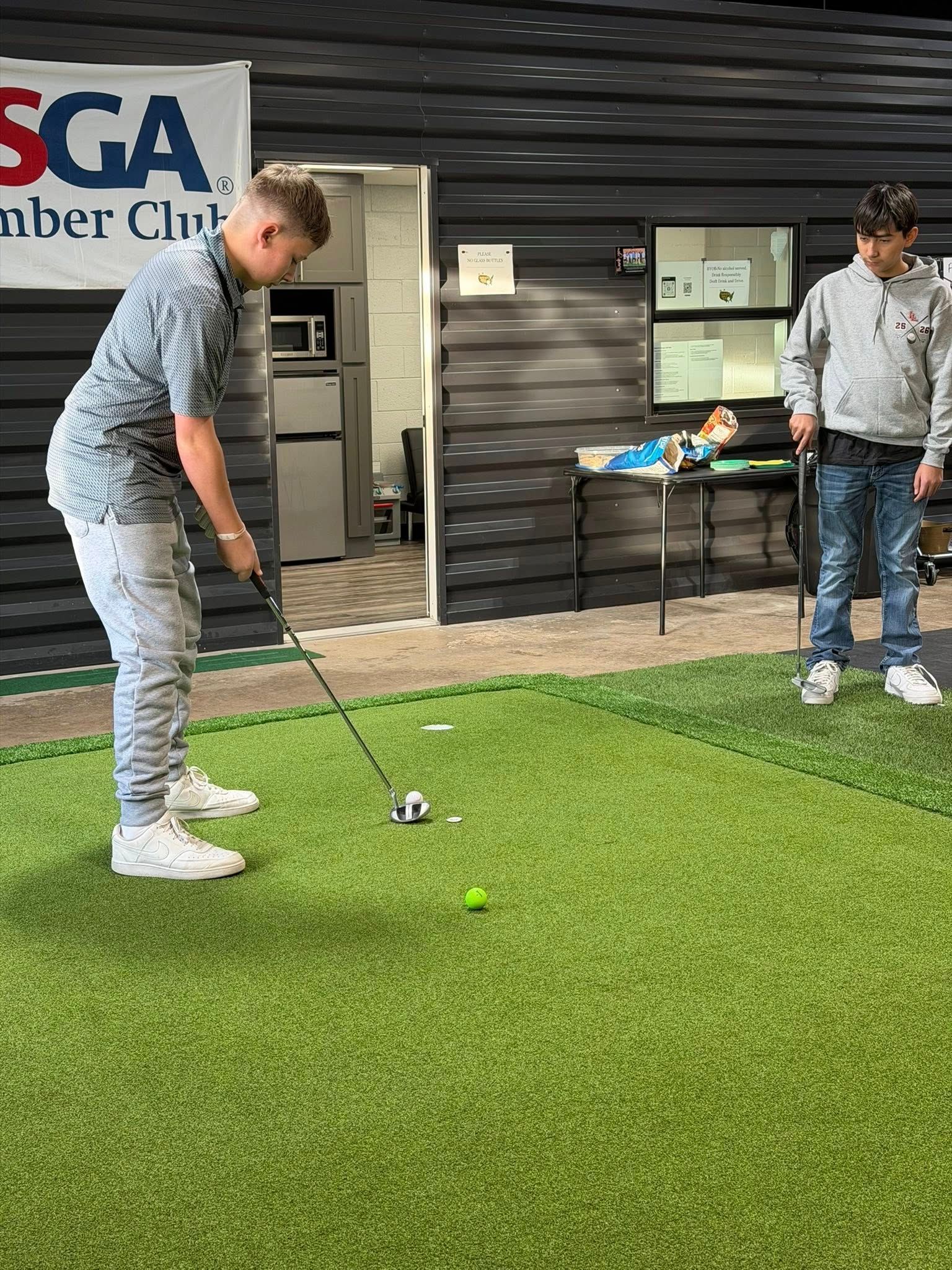 Man putting golf ball on green, another man watches. Indoor golf area.