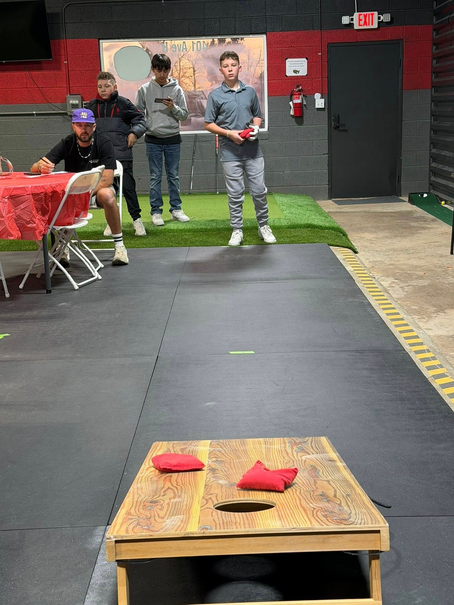 People playing cornhole indoors, throwing beanbags at a wooden board.