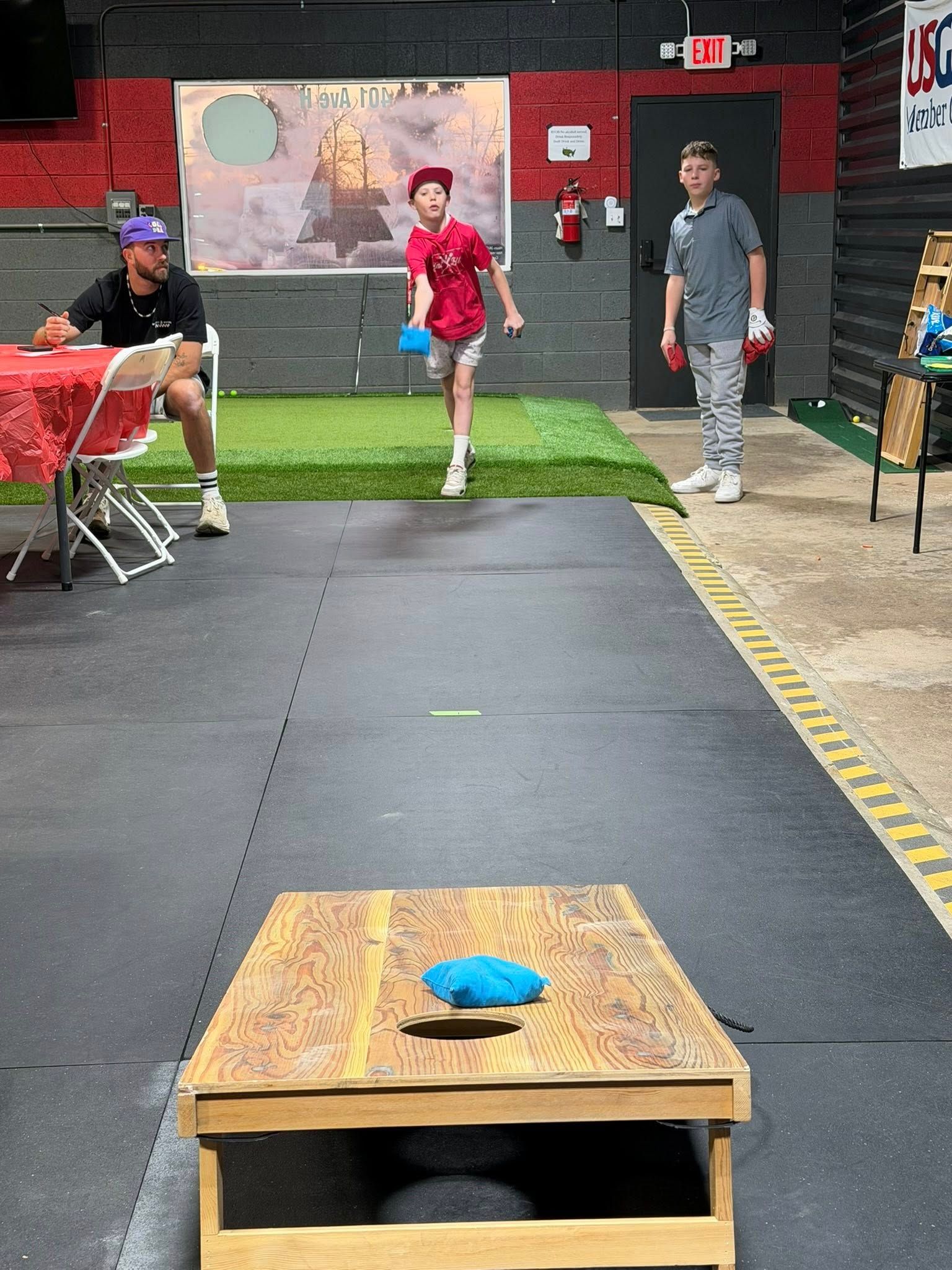Boy throwing a blue bean bag at a cornhole board in an indoor game setting. Two people watching.