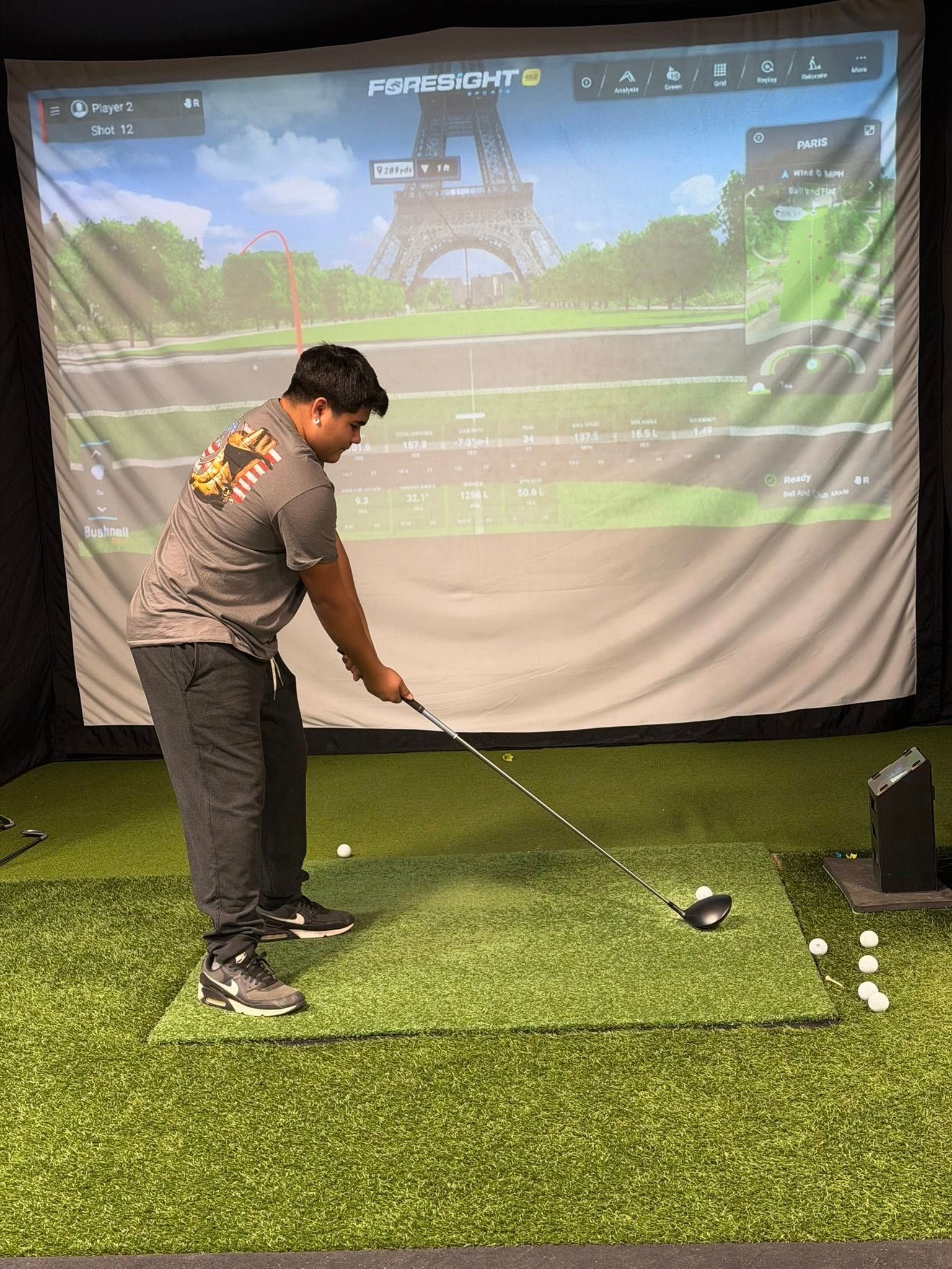 Man swinging a golf club in a golf simulator. He is wearing gray shirt, black pants, black shoes. Eiffel Tower backdrop.