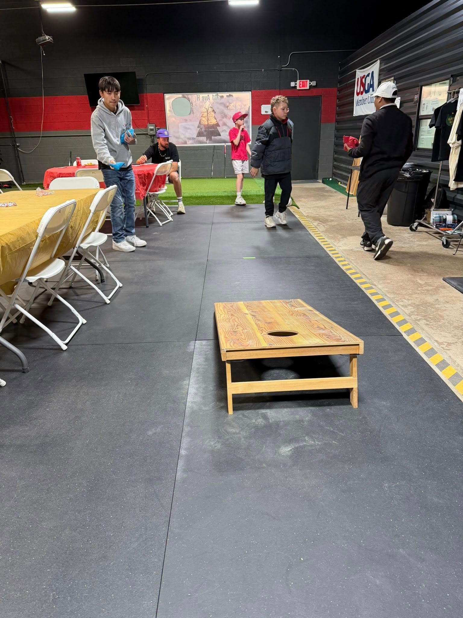 People playing bean bag toss inside a gym; some watching, one about to toss. Table and chairs nearby.