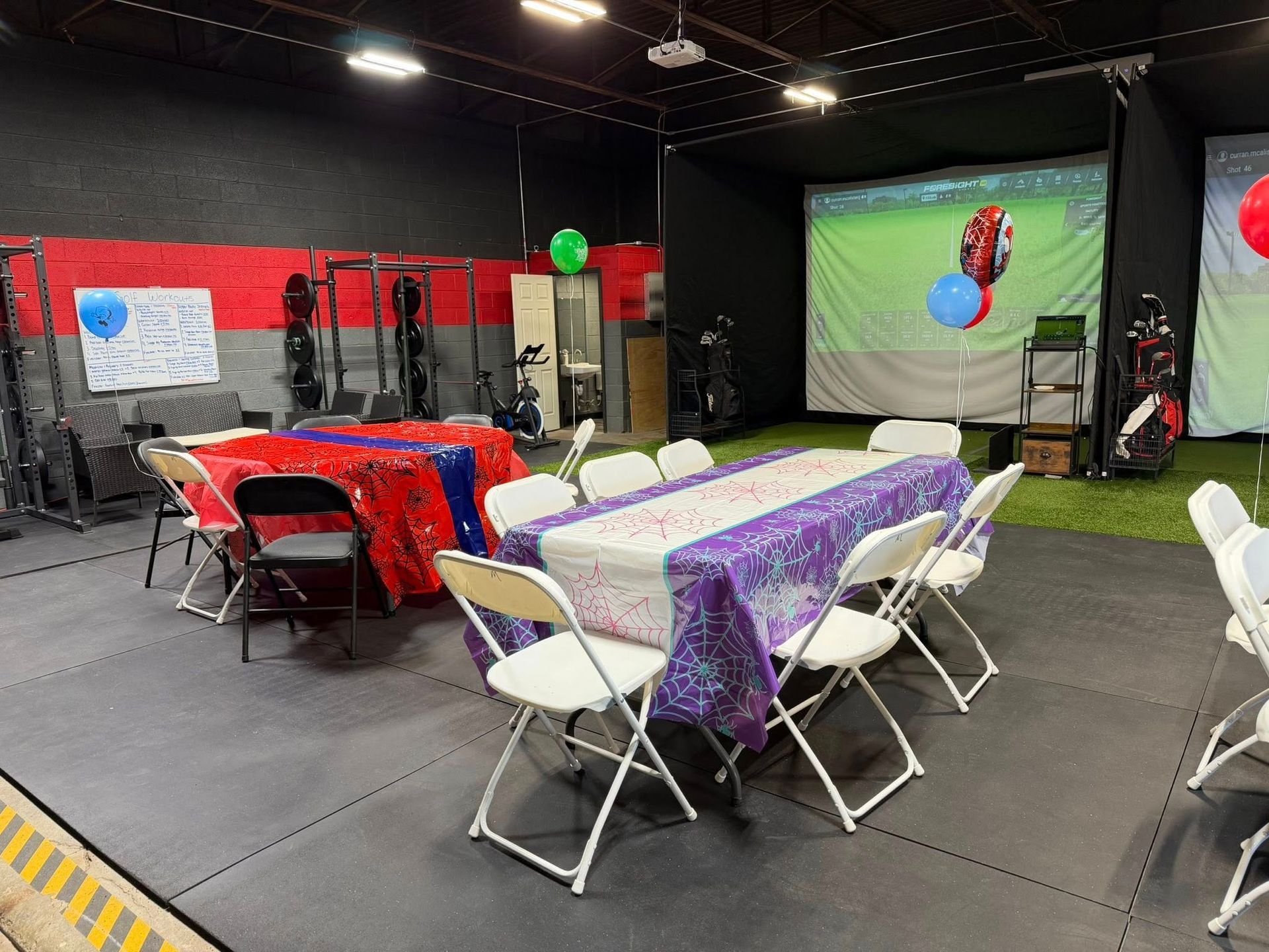 Party tables in a gym with a golf simulator screen, balloons, and weightlifting equipment.