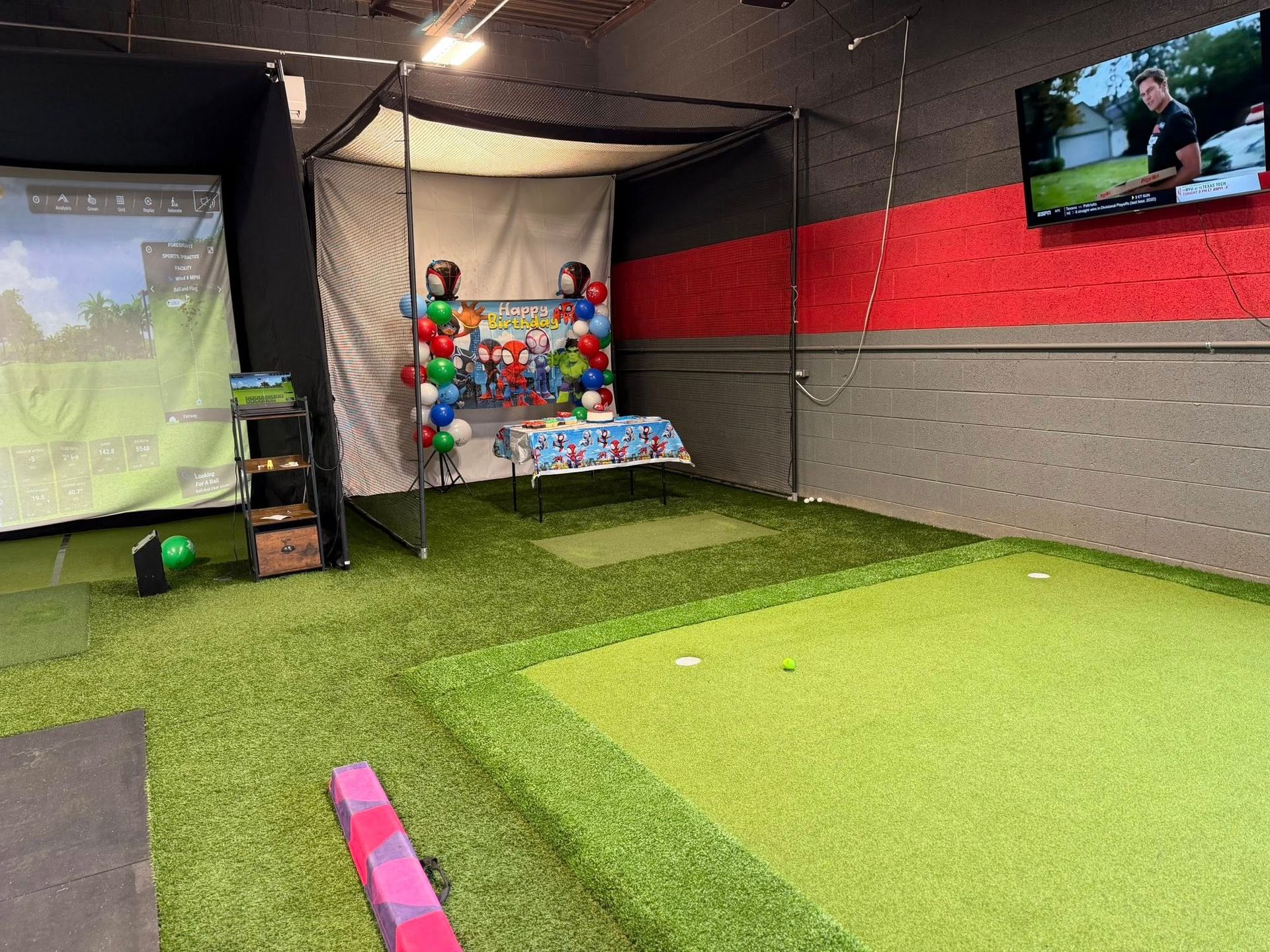 Indoor golf simulator with turf, screen displaying a golf course, and party decorations.