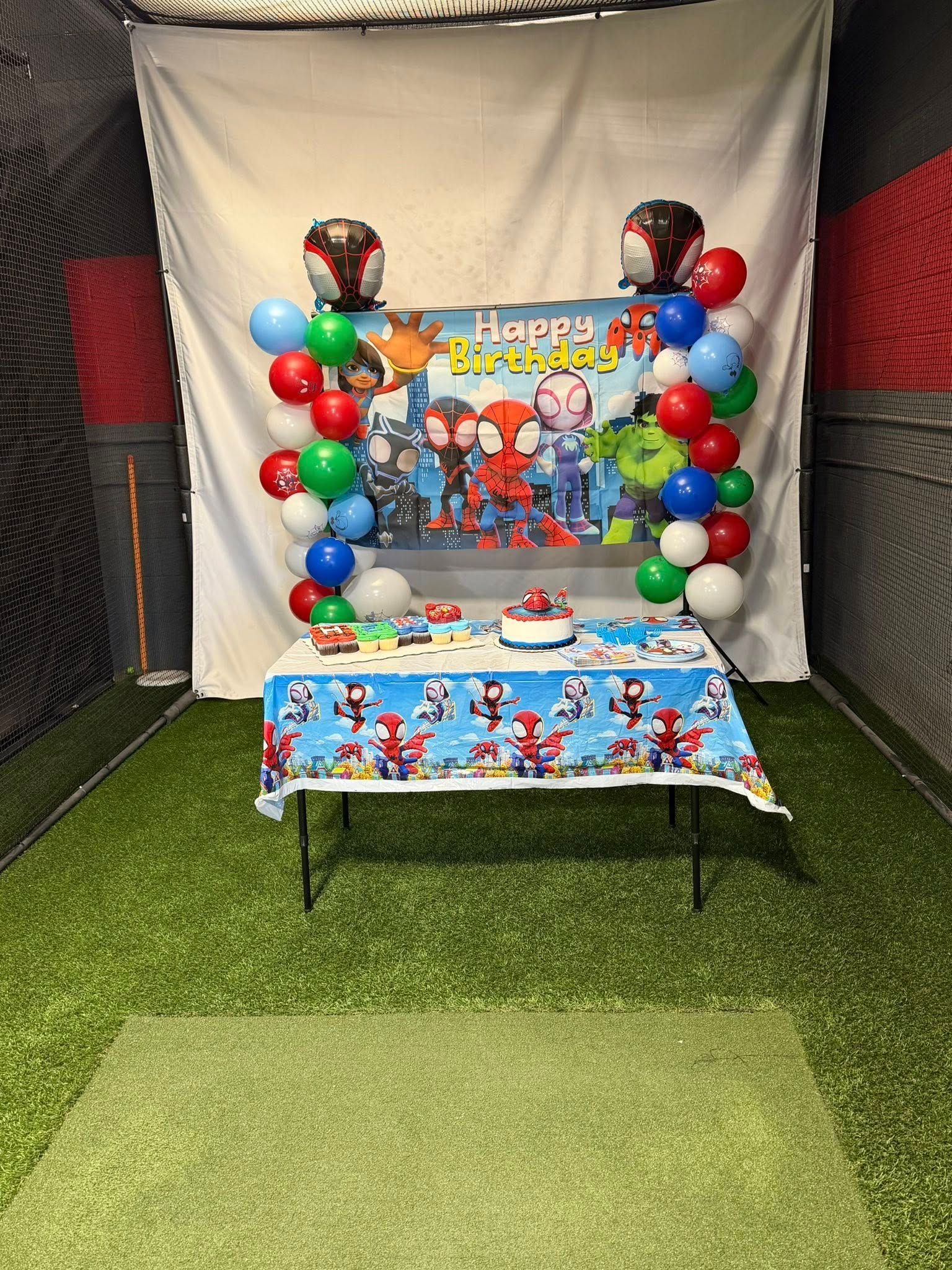 Birthday party table decorated with balloons, a superhero banner, and cake. Set in a room with turf and netting.