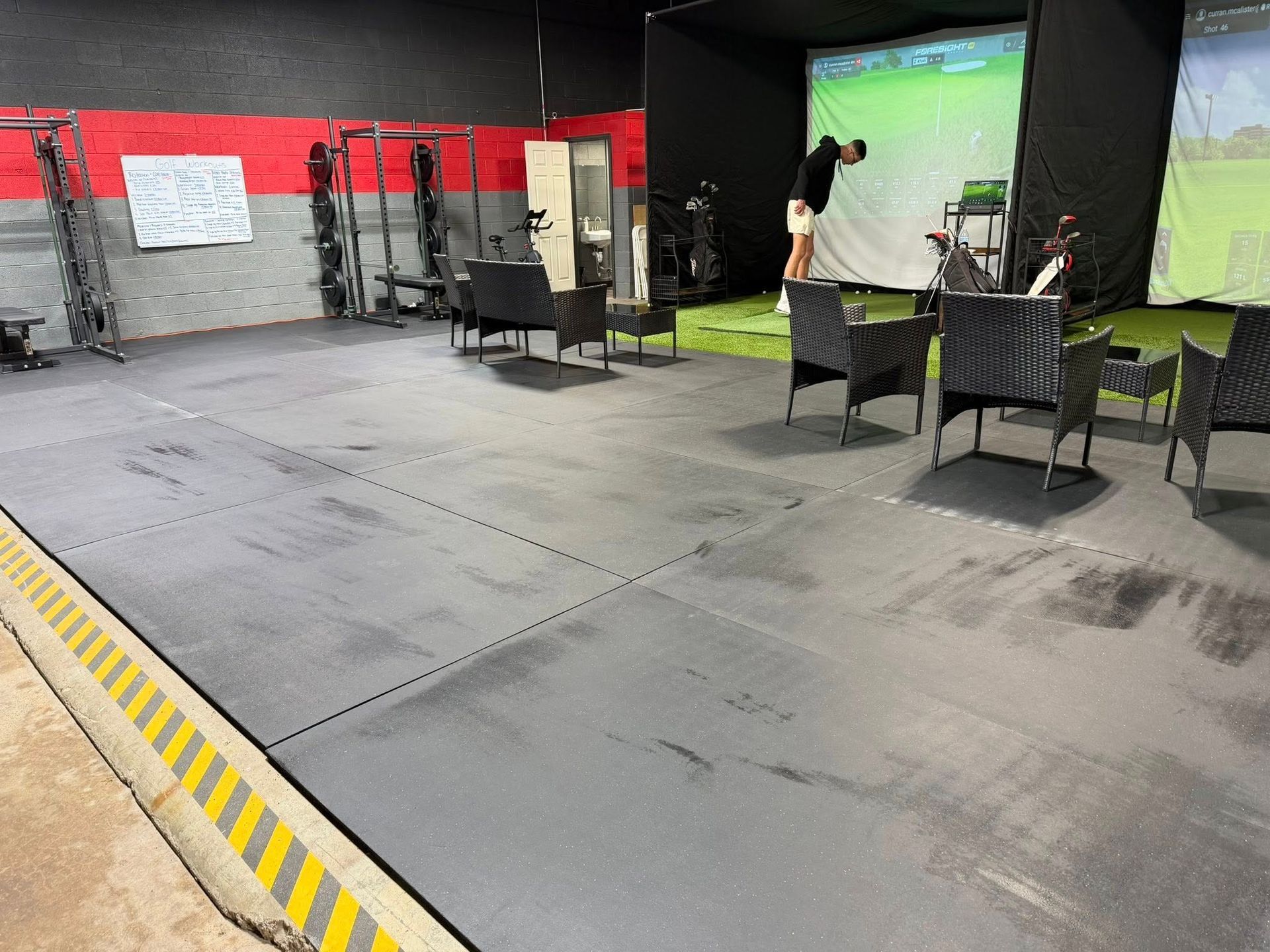 Interior of a golf simulator bay with a person swinging a golf club. Gray floor, black chairs.