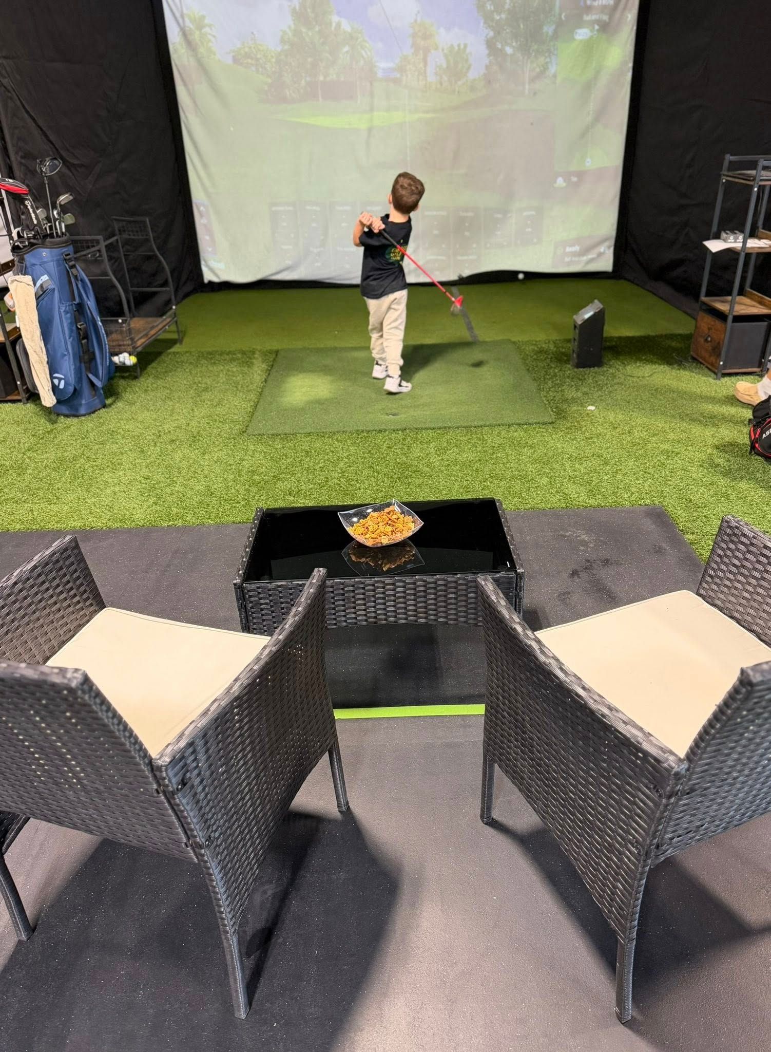 A person swings a golf club in an indoor golf simulator. Two wicker chairs face a low table.