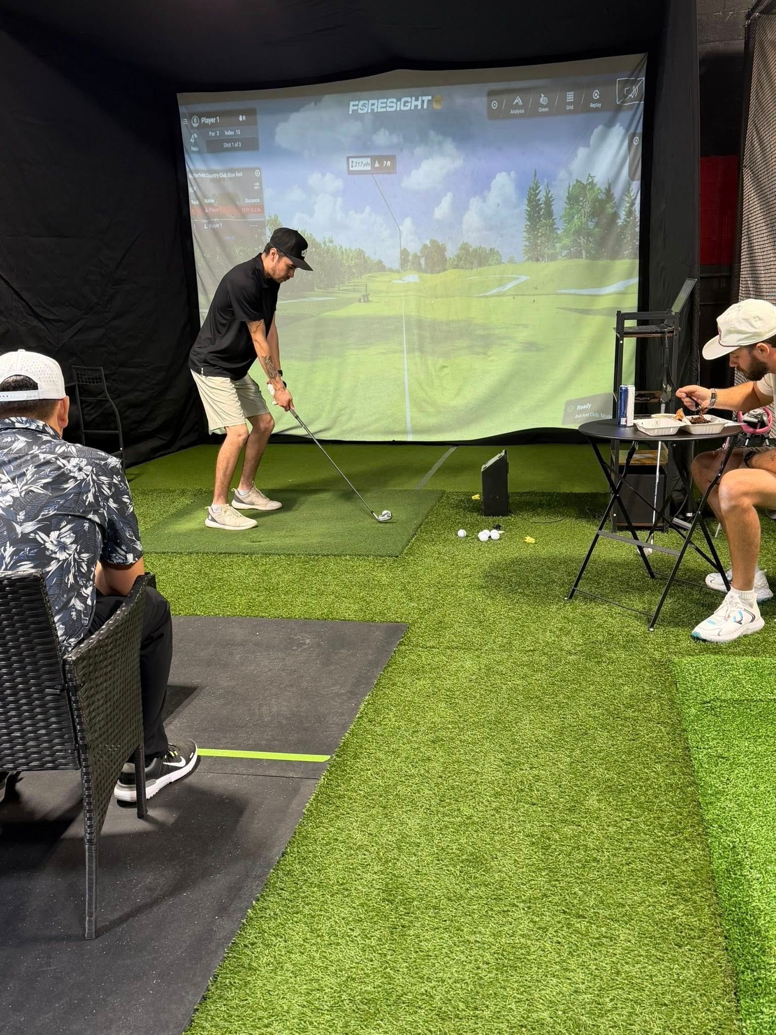 Man swings golf club at simulator screen. Two others watch, one seated at a table. Indoor setting with artificial turf.