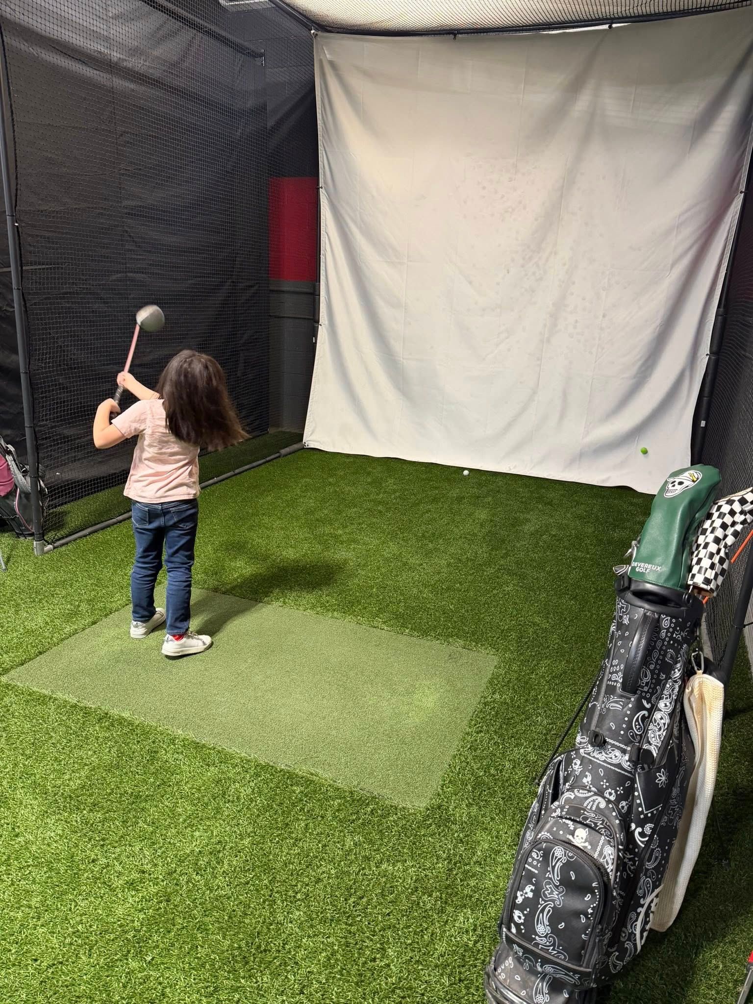 Child in a golf simulator, swinging club at a screen. Green turf, net enclosure, golf bag.