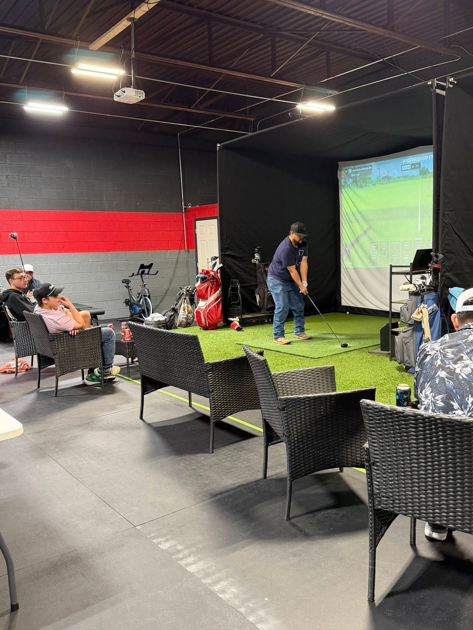 Man swinging a golf club in an indoor golf simulator. People watch from chairs, with equipment and screen visible.