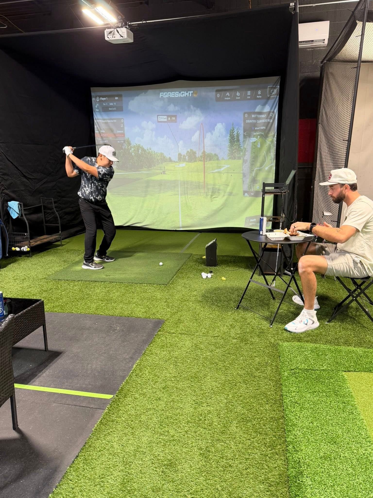 Man swings golf club in indoor simulator, another man watches at a table.