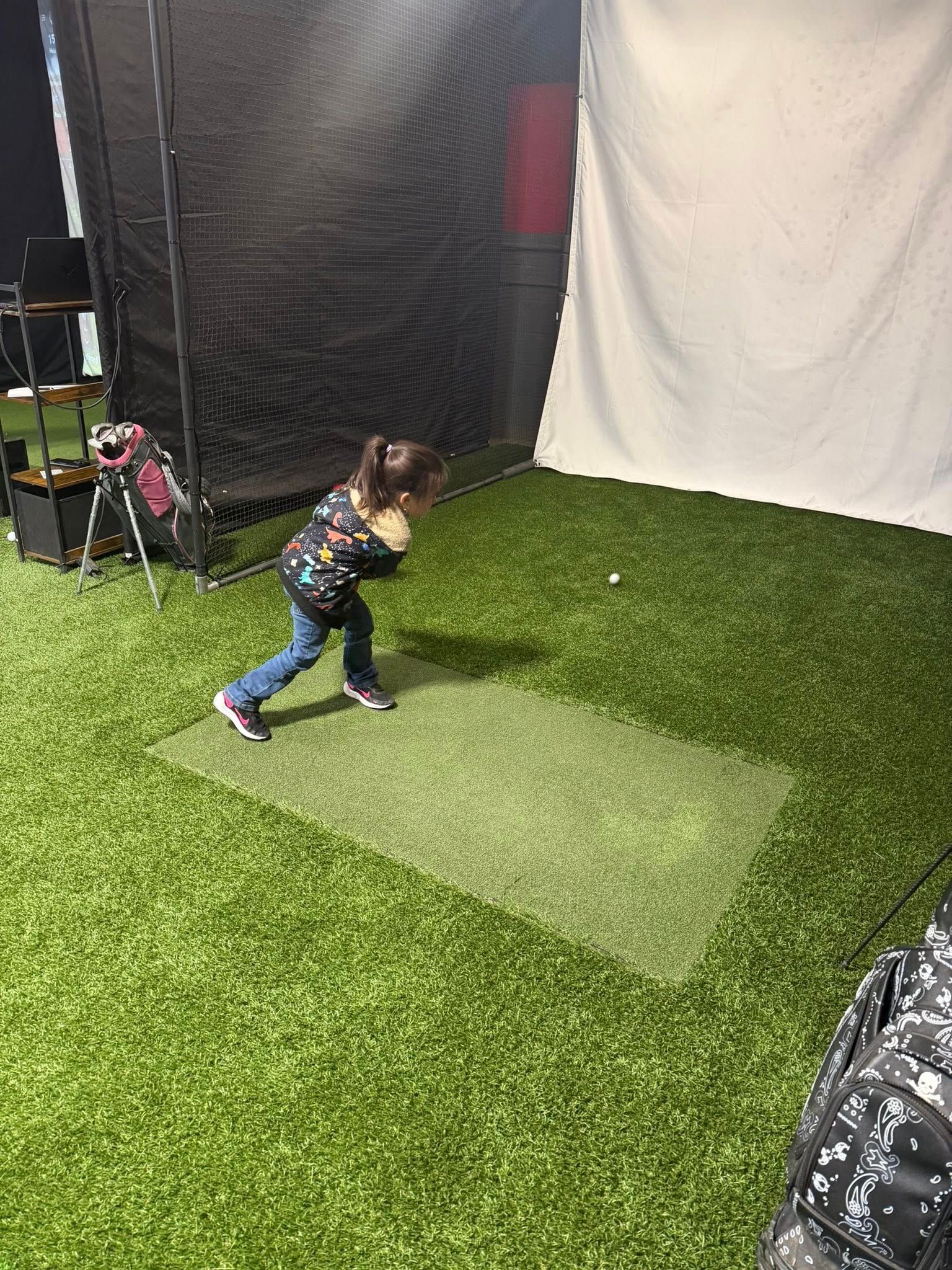 Young child swinging a bat in an indoor batting cage, standing on a green mat.